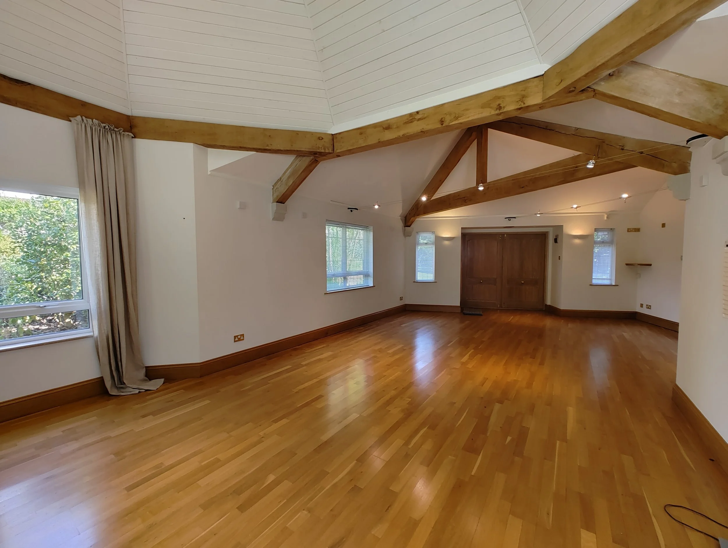 Empty room with hardwood floors, white walls, several windows with blinds, and exposed wooden beams on the ceiling.
