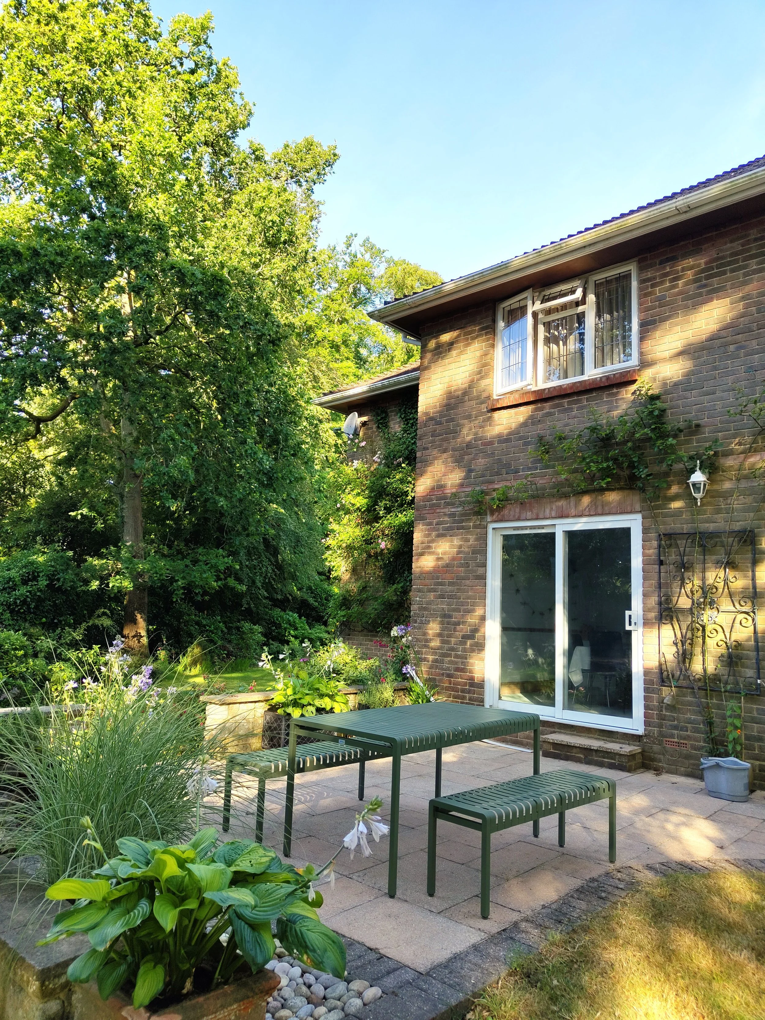 Backyard patio with green benches, surrounded by lush green plants and flowers, adjacent to a brick house with large windows, a small white lantern, and a garden bed.
