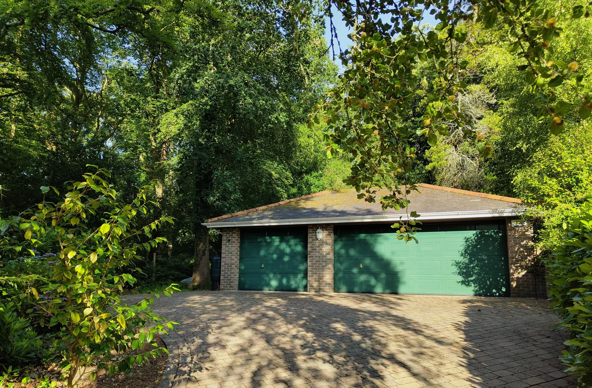 A brick house with green garage doors under a leafy tree canopy. The driveway is paved with bricks, and shadows are cast by the trees.