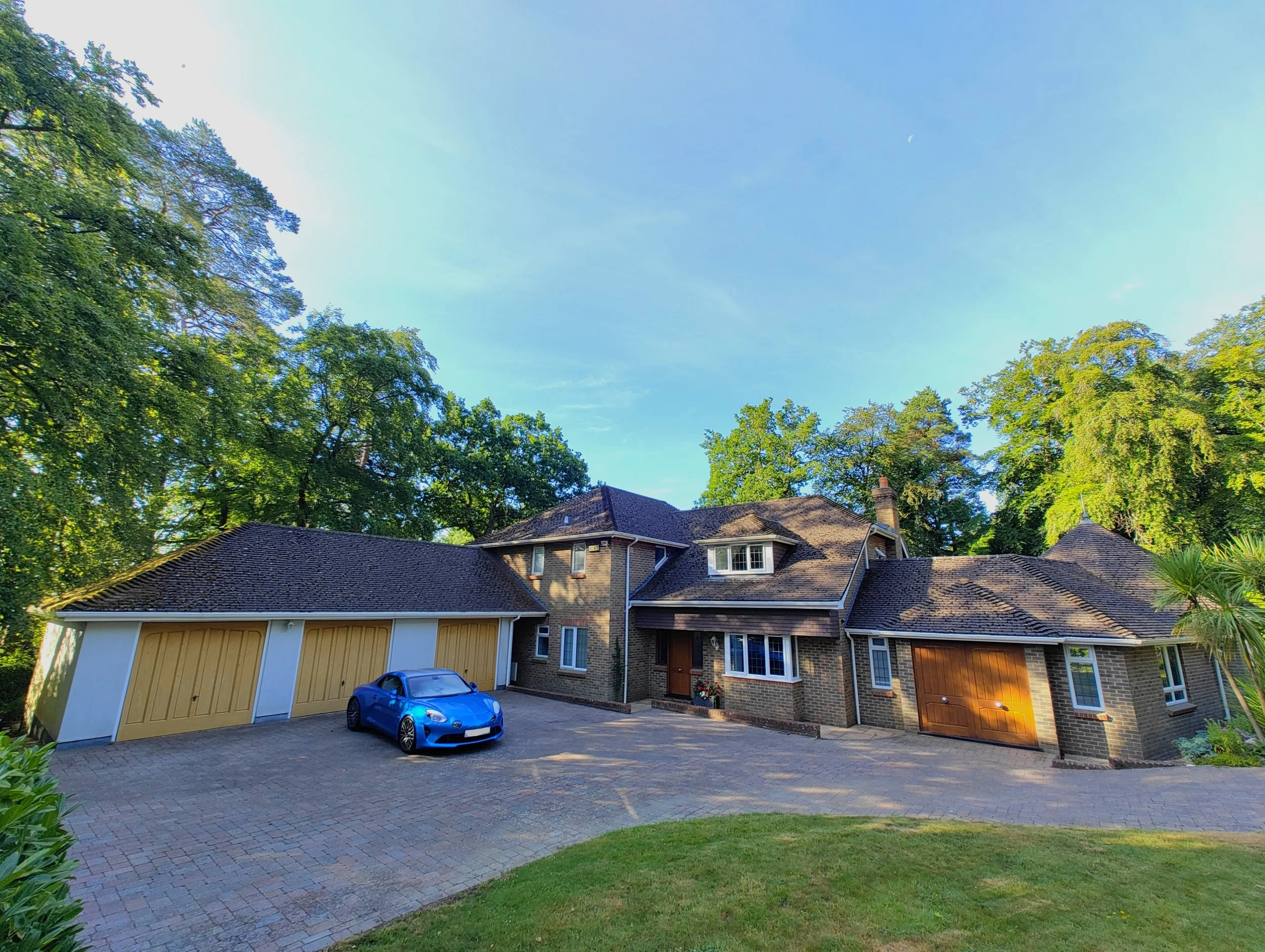 A suburban house with a brick facade, multiple garages, and a blue sports car parked in the driveway, surrounded by tall green trees and a well-kept lawn.
