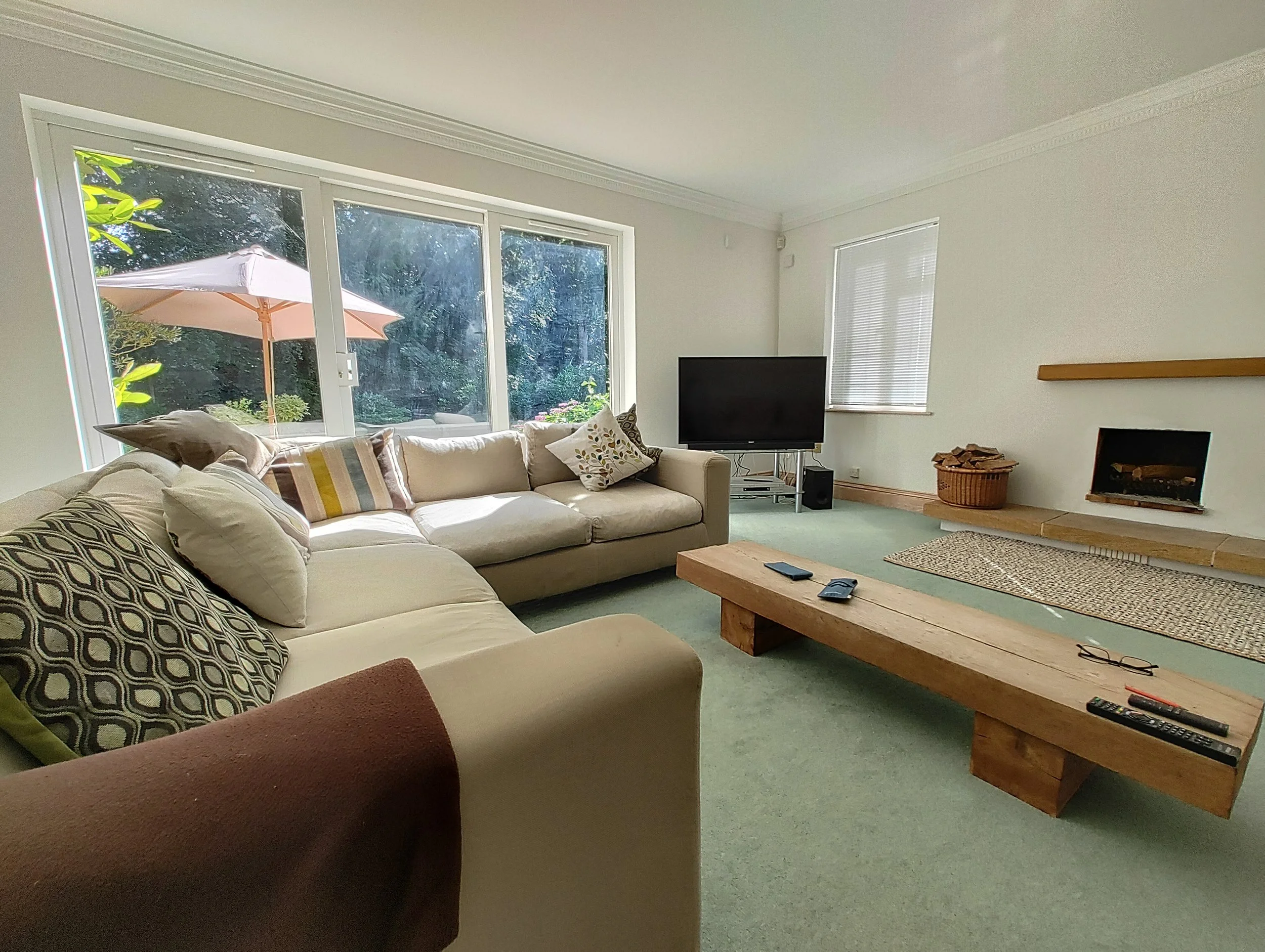 Living room with large windows, beige sofa with patterned pillows, a television on a stand, a wooden coffee table with remote controls and glasses, and a fireplace with a basket of logs, with a view of a patio with an umbrella outside.