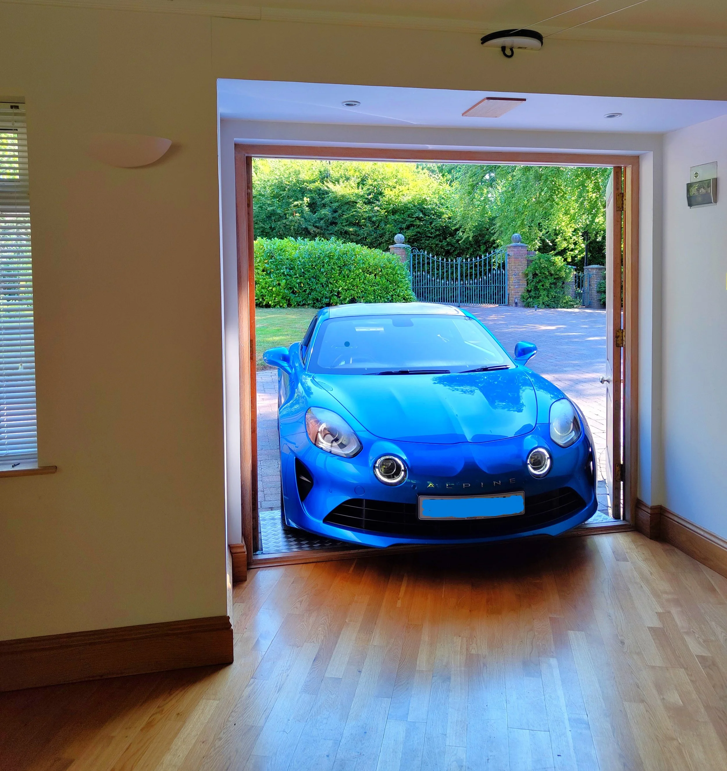 A bright blue sports car parked inside a house garage, visible through an open wooden door. The garage opens to a driveway surrounded by green shrubs and trees.