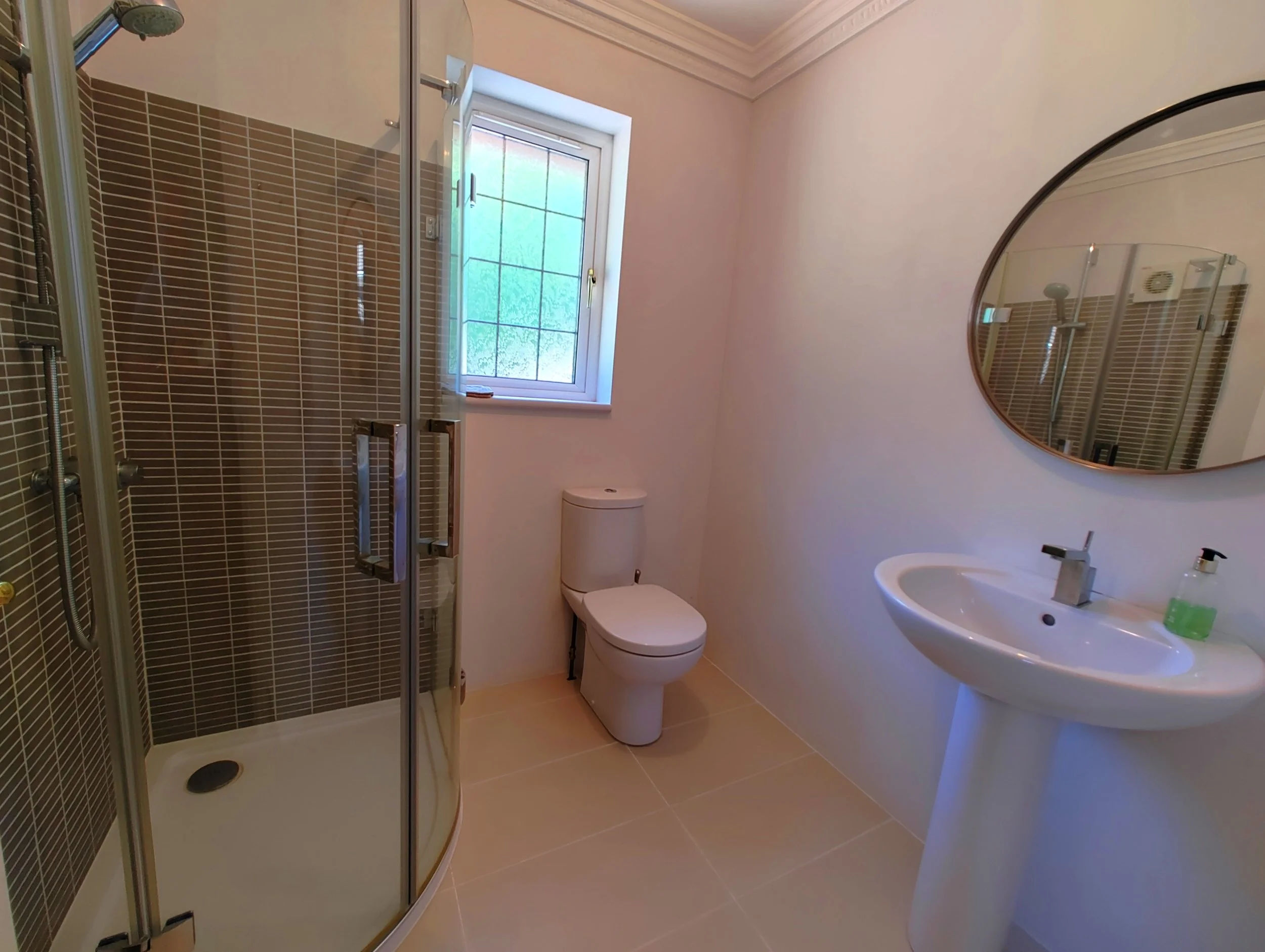 A bathroom with a walk-in shower, a toilet, a sink, a round mirror, and a window with frosted glass, featuring neutral-colored tiles and light-colored walls.