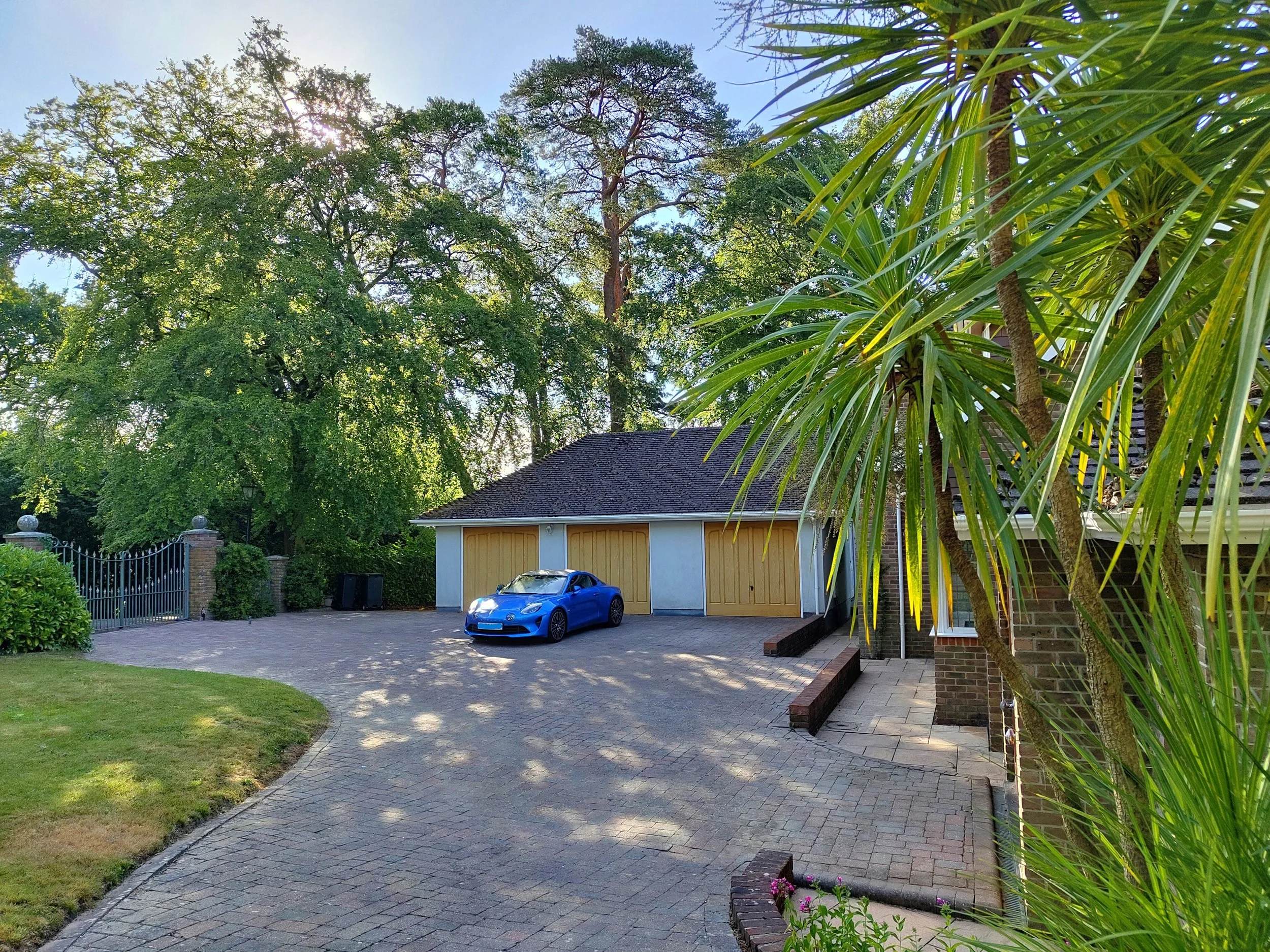 A residential driveway with a blue car parked in front of a garage with tan doors. The driveway is paved with bricks and surrounded by green grass and plants. Tall trees and a house are visible in the background, with sunlight filtering through the l