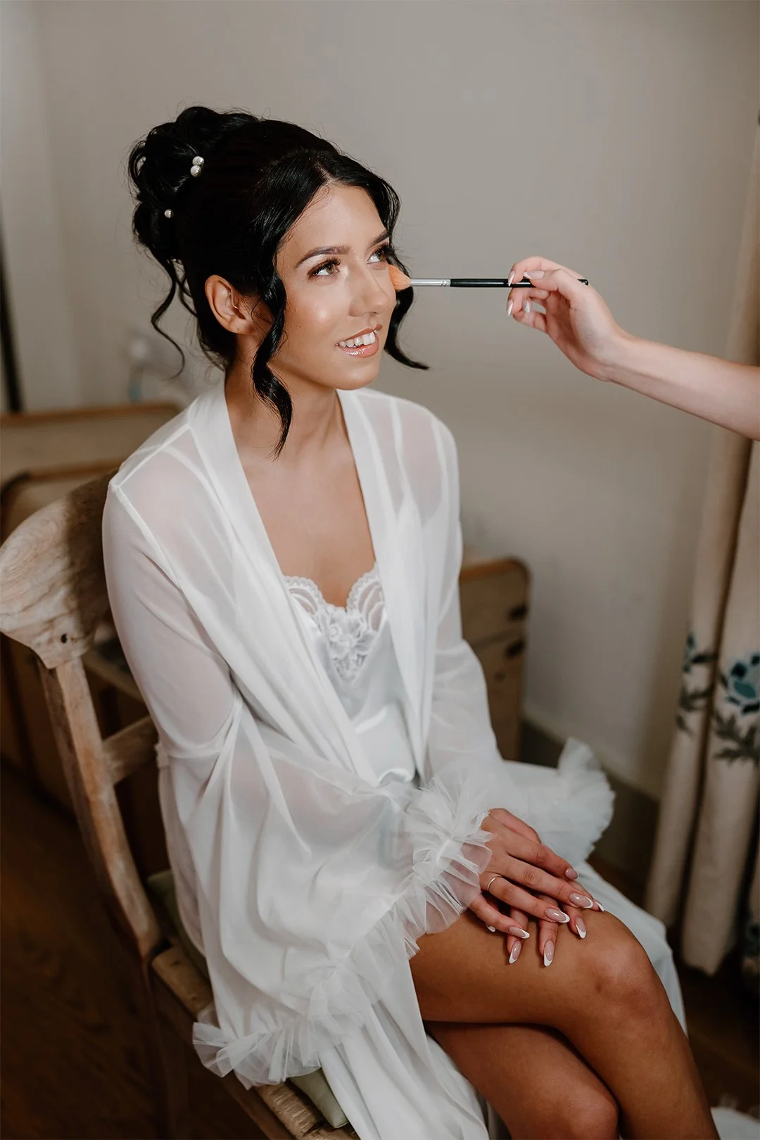 A woman with dark hair styled in an elegant updo with pearl accents is sitting on a wooden chair, smiling as a makeup artist applies makeup to her nose with a brush. She is wearing a white satin robe with ruffled cuffs and lace detailing. She has a neutral, soft expression and is looking slightly upward.