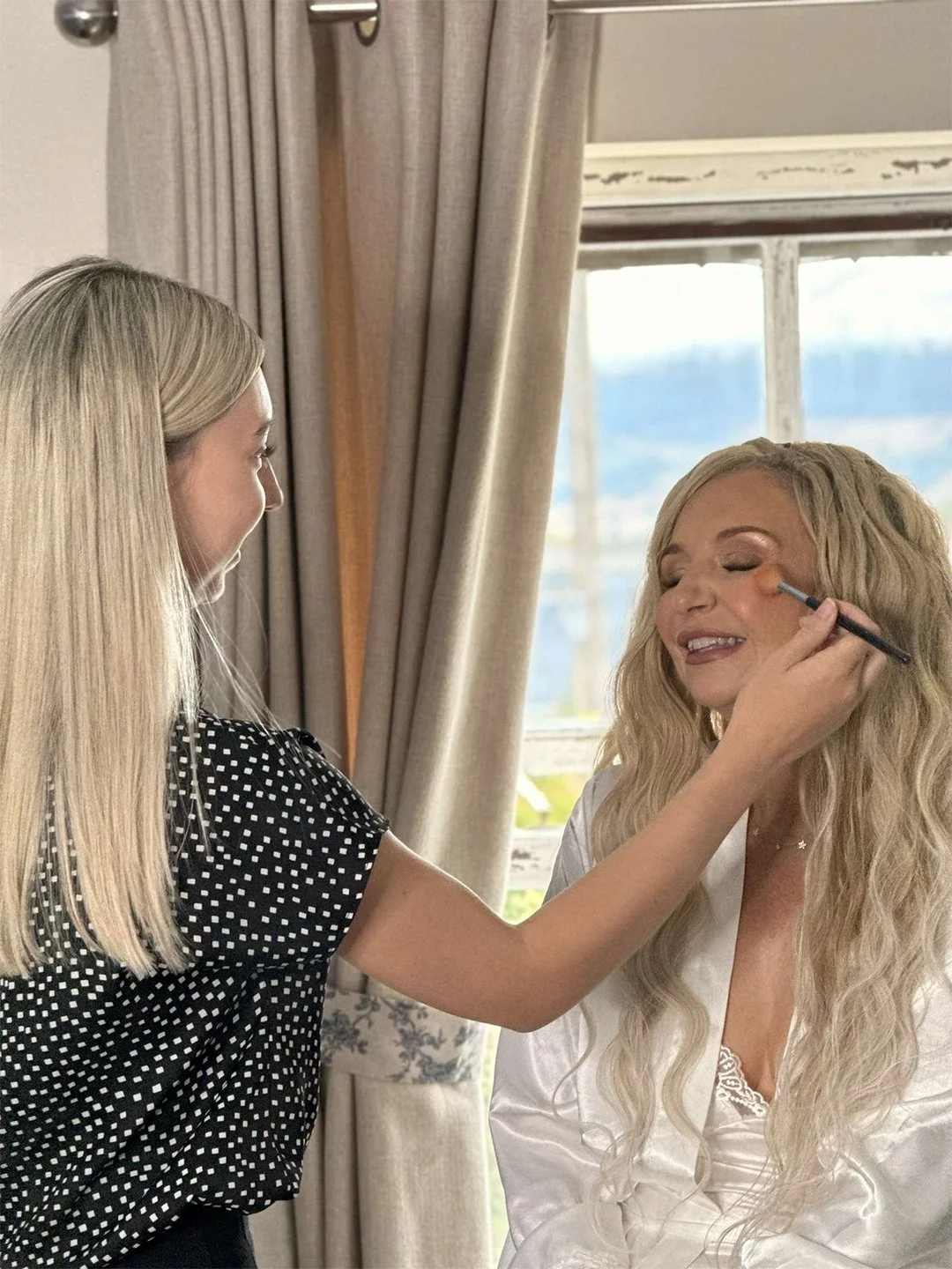 A woman applying makeup to another woman near a window with beige curtains.