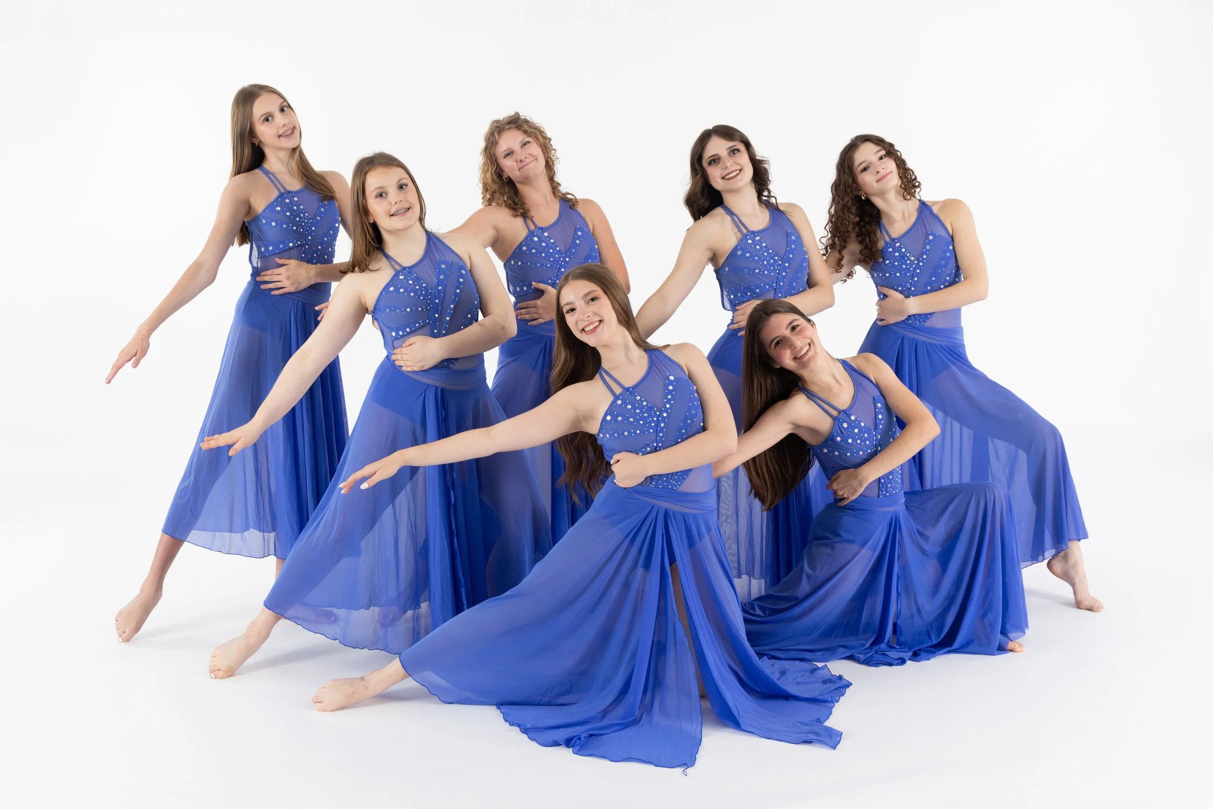 Group of seven young girls in matching blue dresses posing in a dance studio with a white background.