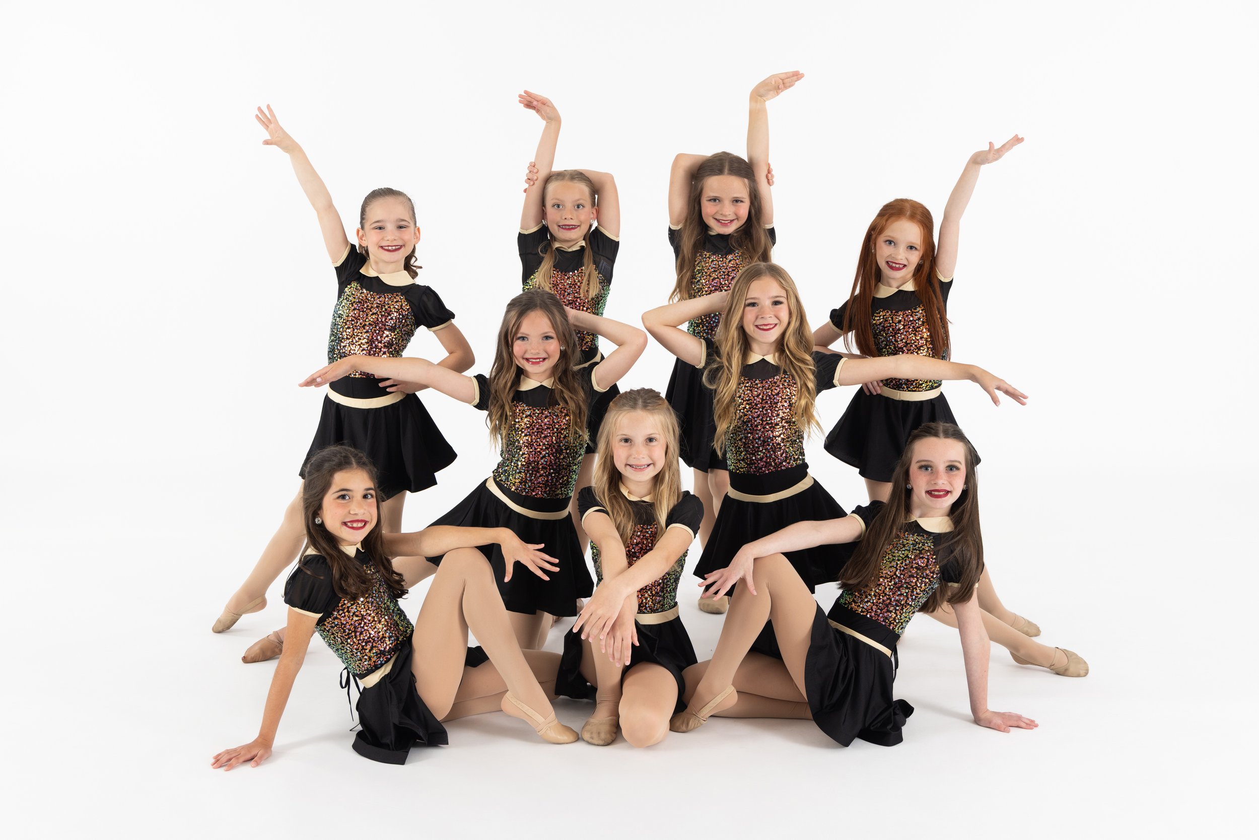 Group of seven young girls dressed in black and glittery costumes, posing in a dance formation against a white background.