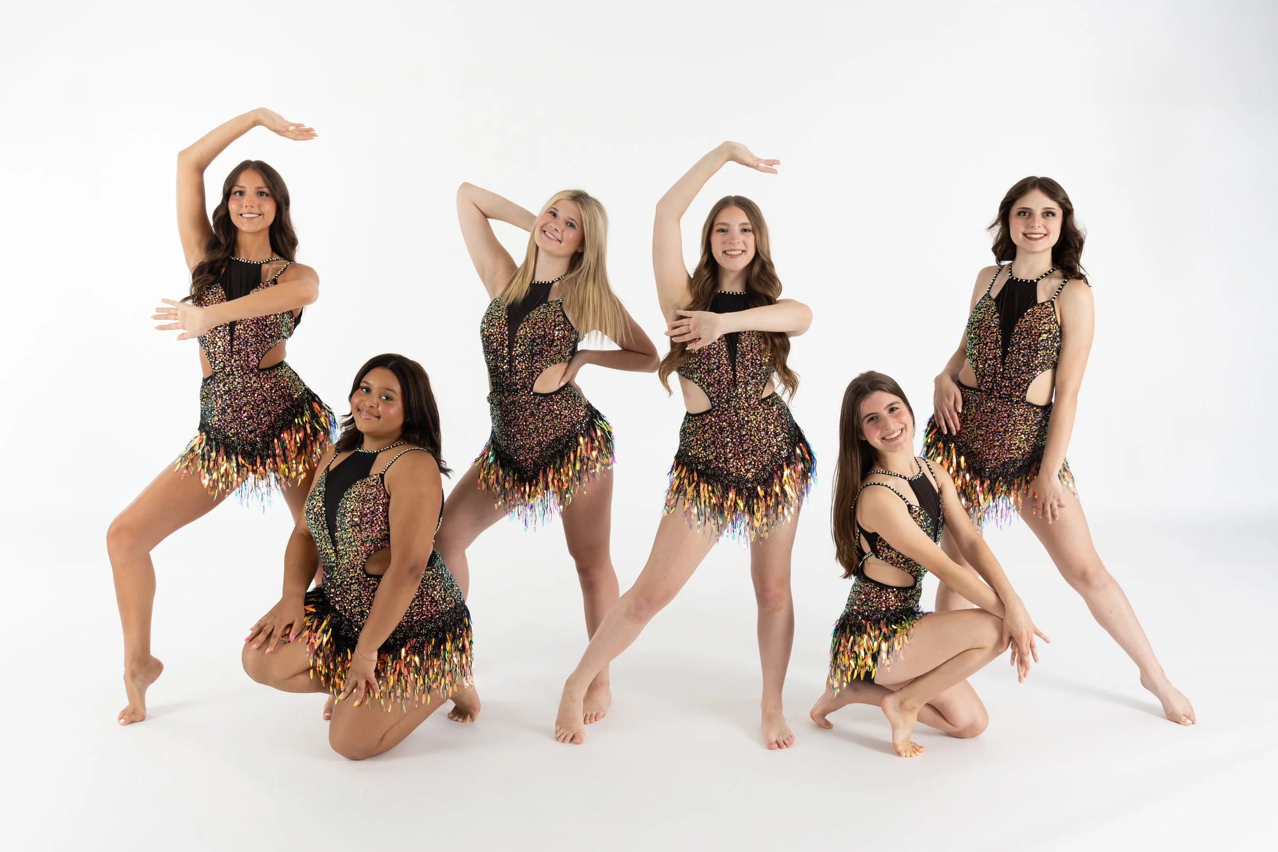 A group of seven young women dressed in matching colorful, sequin, and feather dance costumes posing in a studio with a white background. Five women are standing, and two are sitting or kneeling, all smiling.
