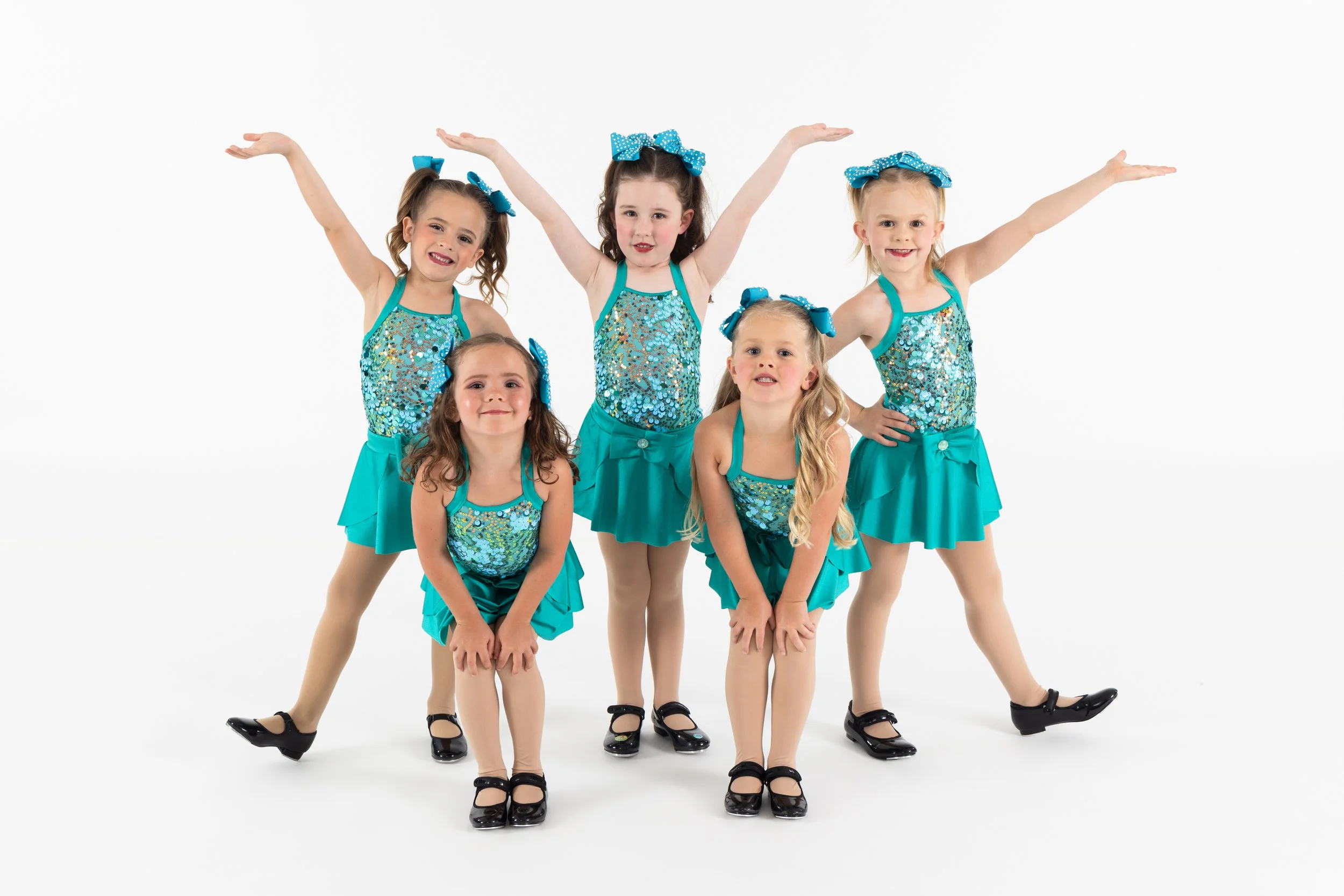 Five young girls dressed in matching turquoise sparkly dance costumes with bows in their hair, standing in a formation with some girls crouching and others extending their arms and legs.