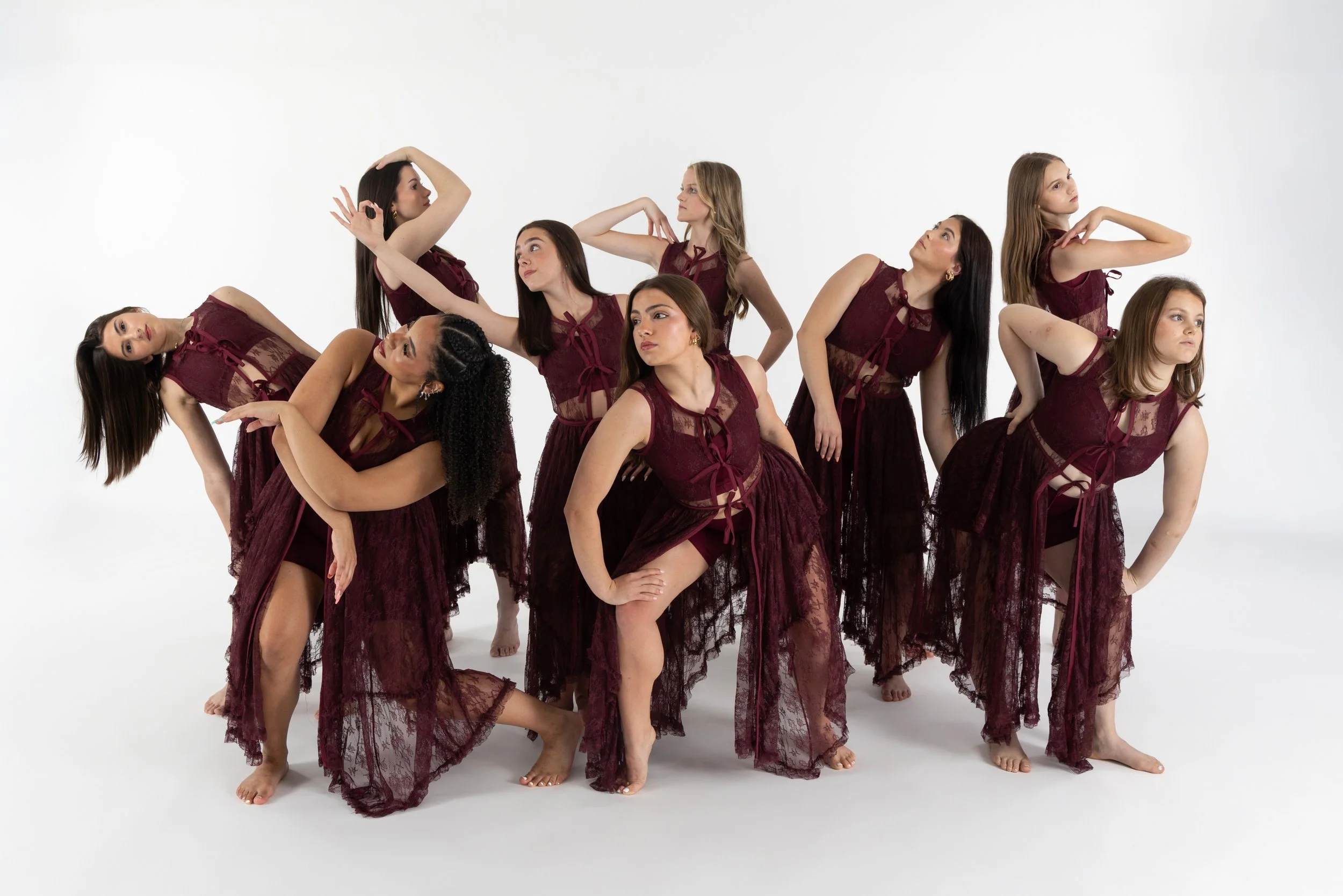 A group of nine women in matching burgundy lace dresses posing in a white studio. They are arranged in a dynamic formation, with some leaning, standing, or sitting, and exhibiting various contemplative and artistic poses.