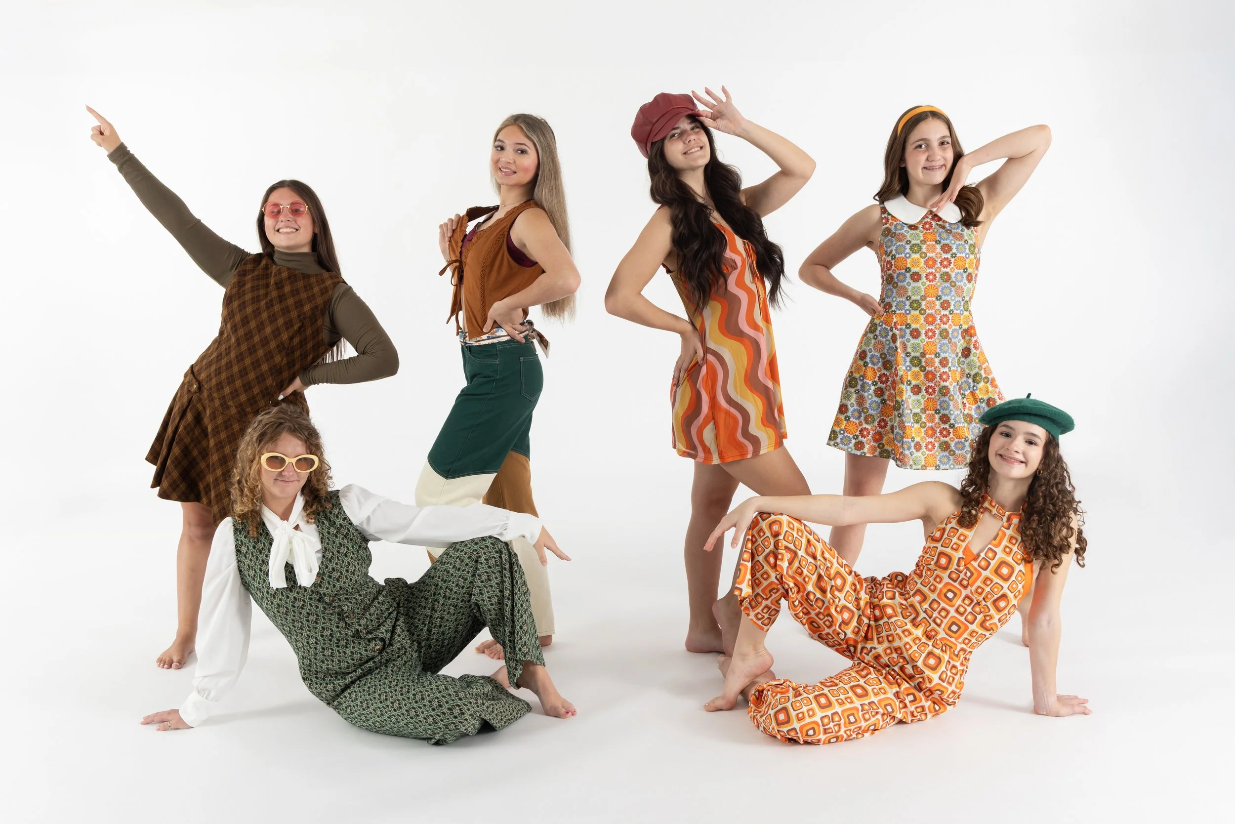 Group of seven young women posing playfully against a white background, wearing colorful, vintage-inspired outfits.