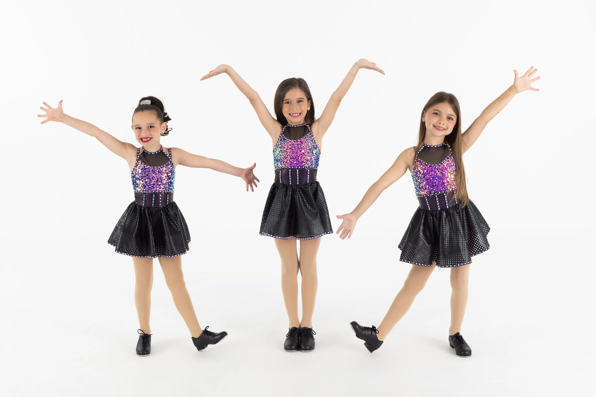 Three young girls in dance costumes pose with arms raised and smiling in front of a white background.