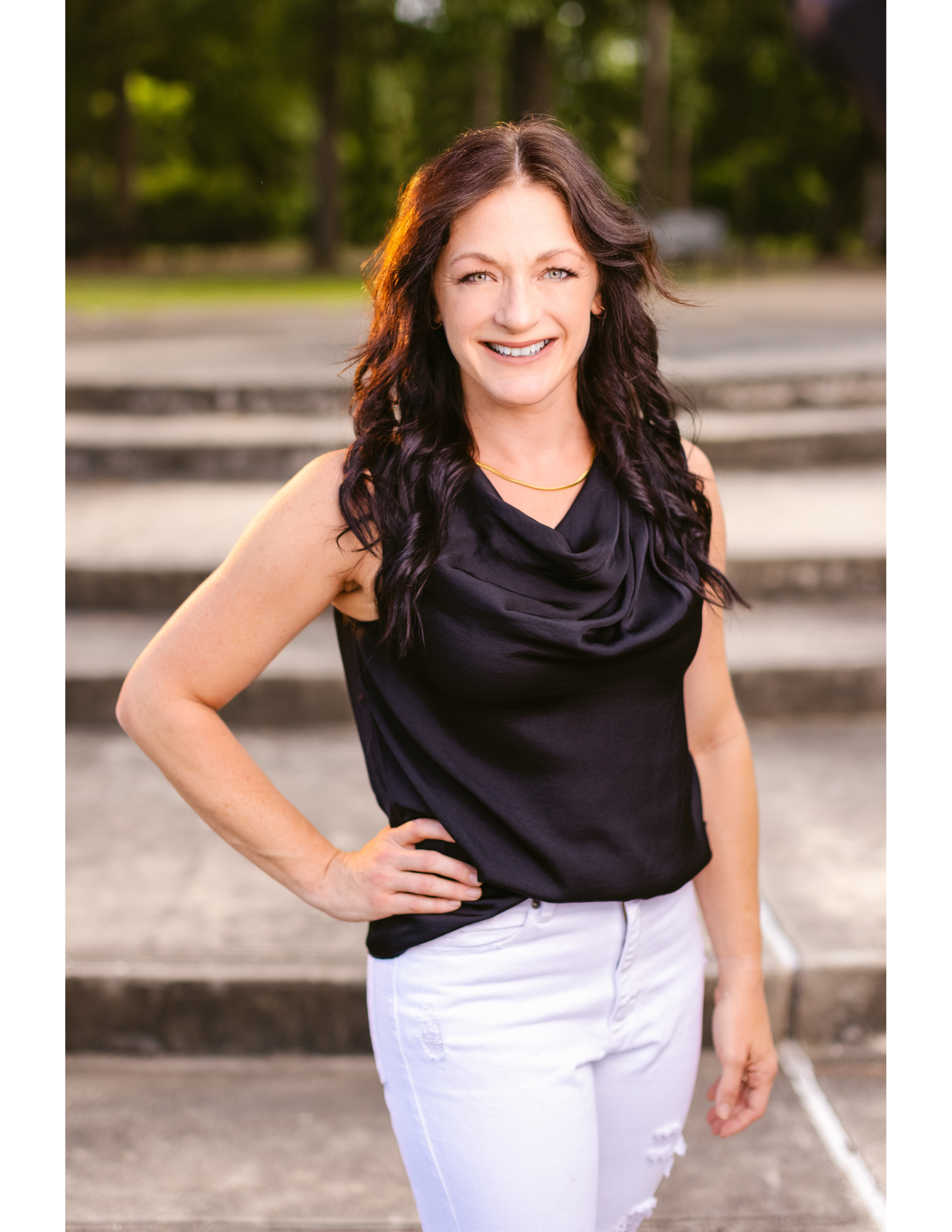 A woman with dark wavy hair smiling outdoors, wearing a black top, white jeans with ripped knee, and a gold necklace, standing with her right hand on her hip.