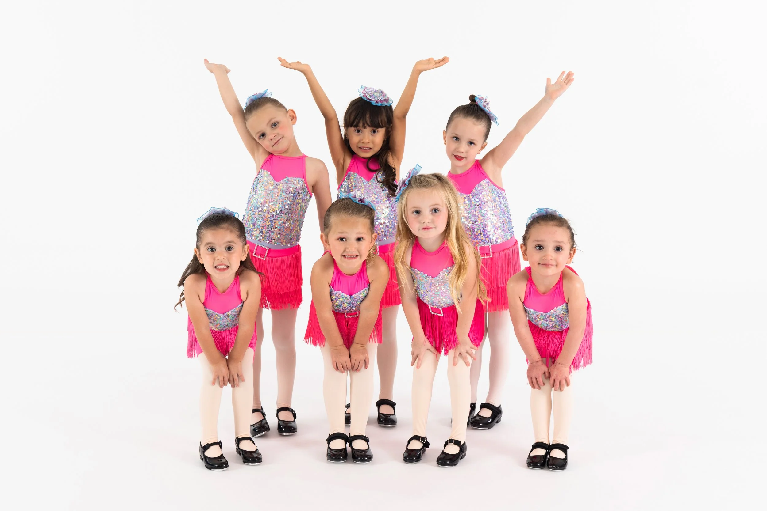 Seven young girls in matching pink and sparkly costumes standing on a white background, some with arms raised or bent at the elbows, in a dance or gymnastics pose.
