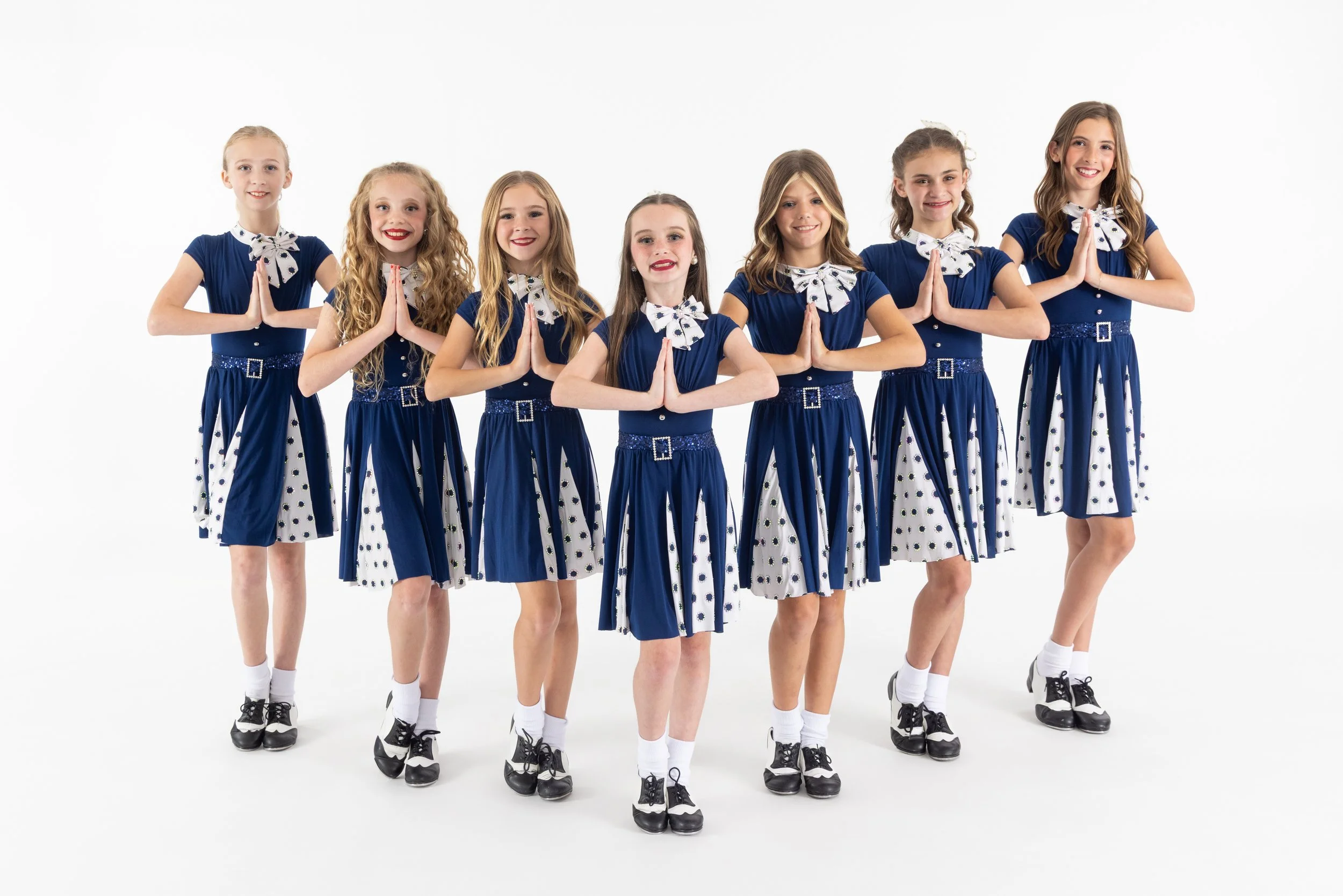 Seven young girls dressed in blue and white cheerleading uniforms, standing in a row with hands pressed together in prayer-like gesture, on a white background.