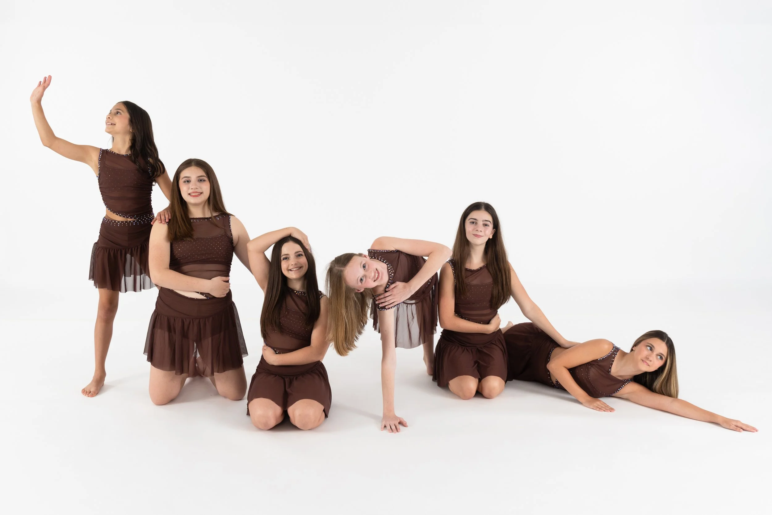 Group of seven young girls wearing matching brown dresses with sheer fabric and embellishments, posing in various positions on a white background.