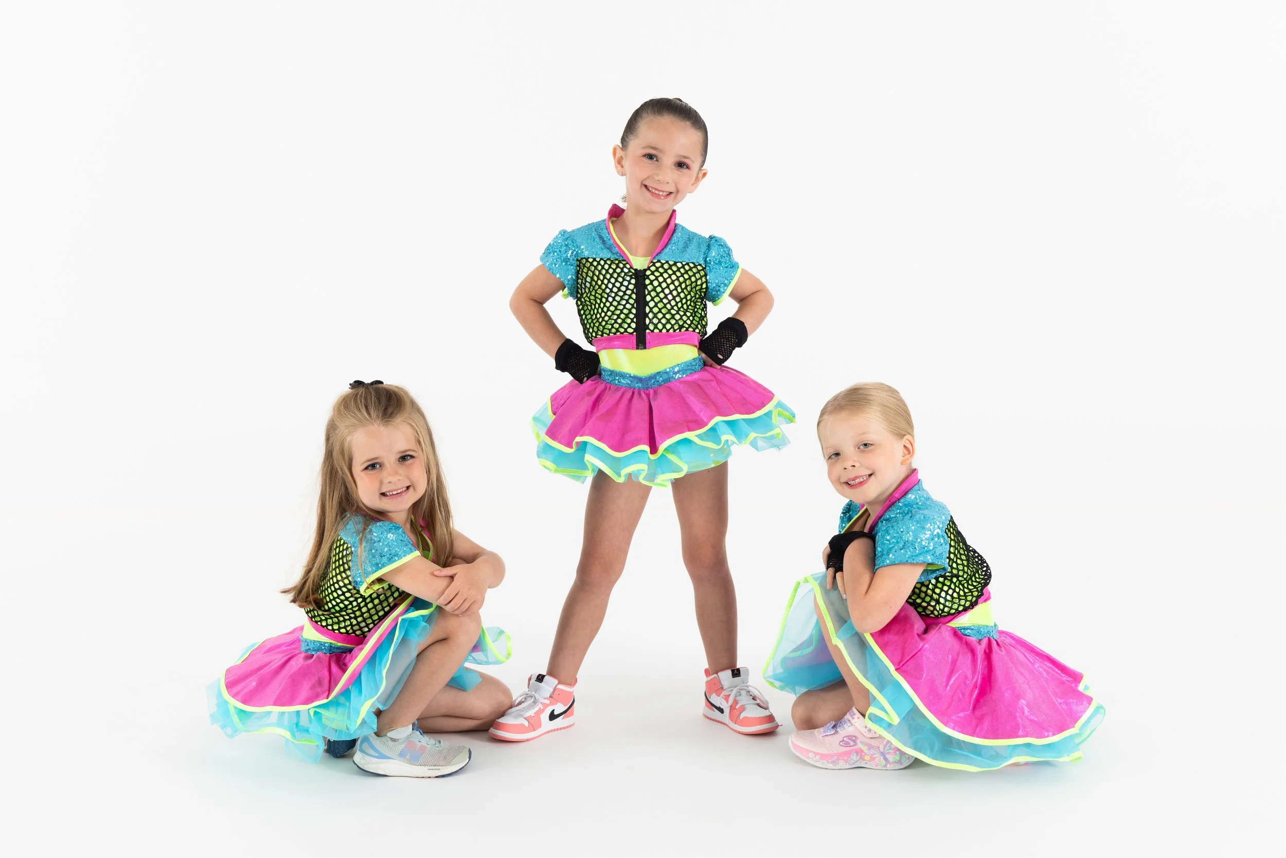 Four young girls dressed in colorful dance costumes with pink, blue, green, and yellow accents posing together against a white background.