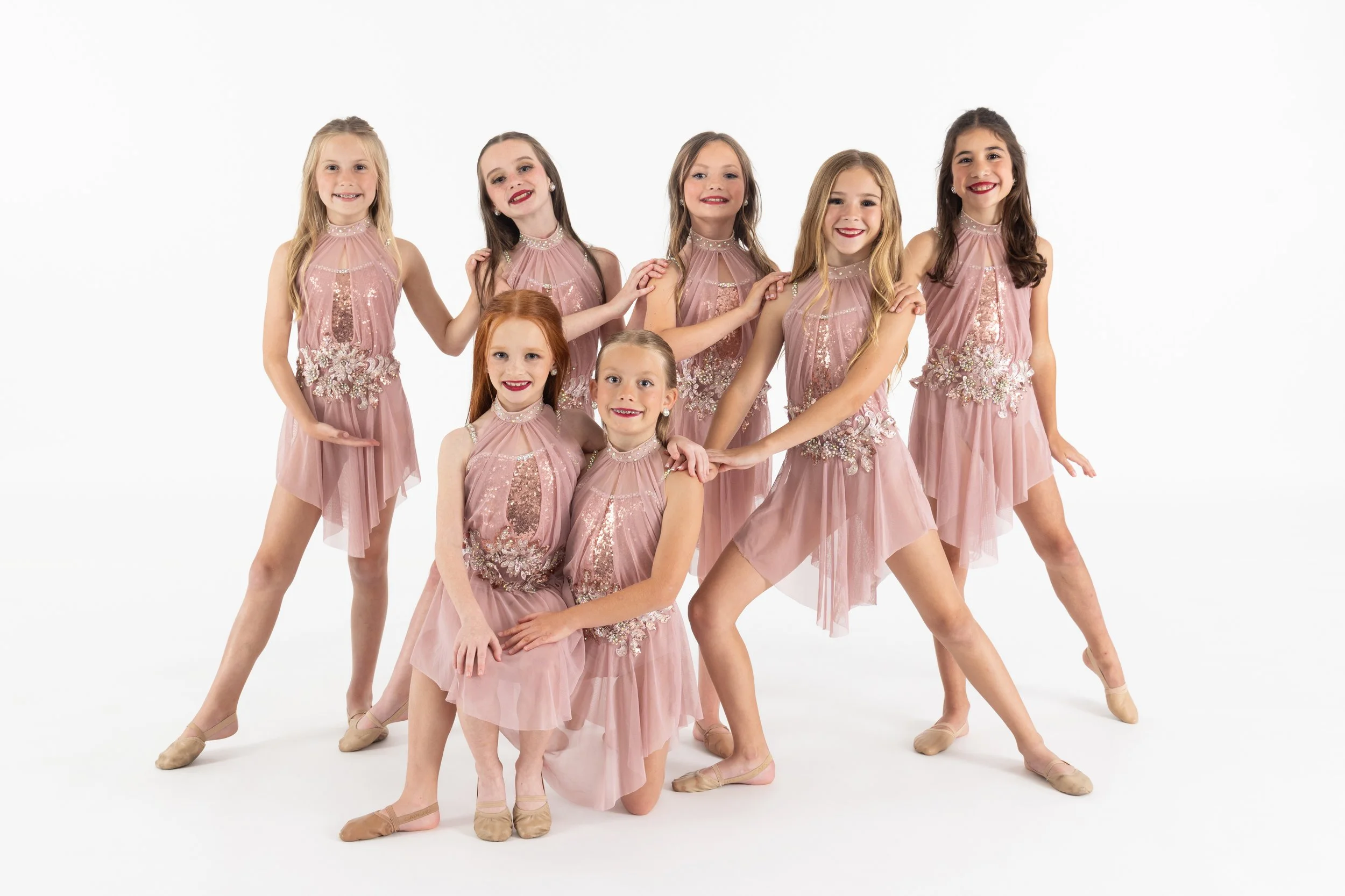 Group of eight young girls in pink ballet costumes standing together, some smiling, posing in dance studio with plain white background.