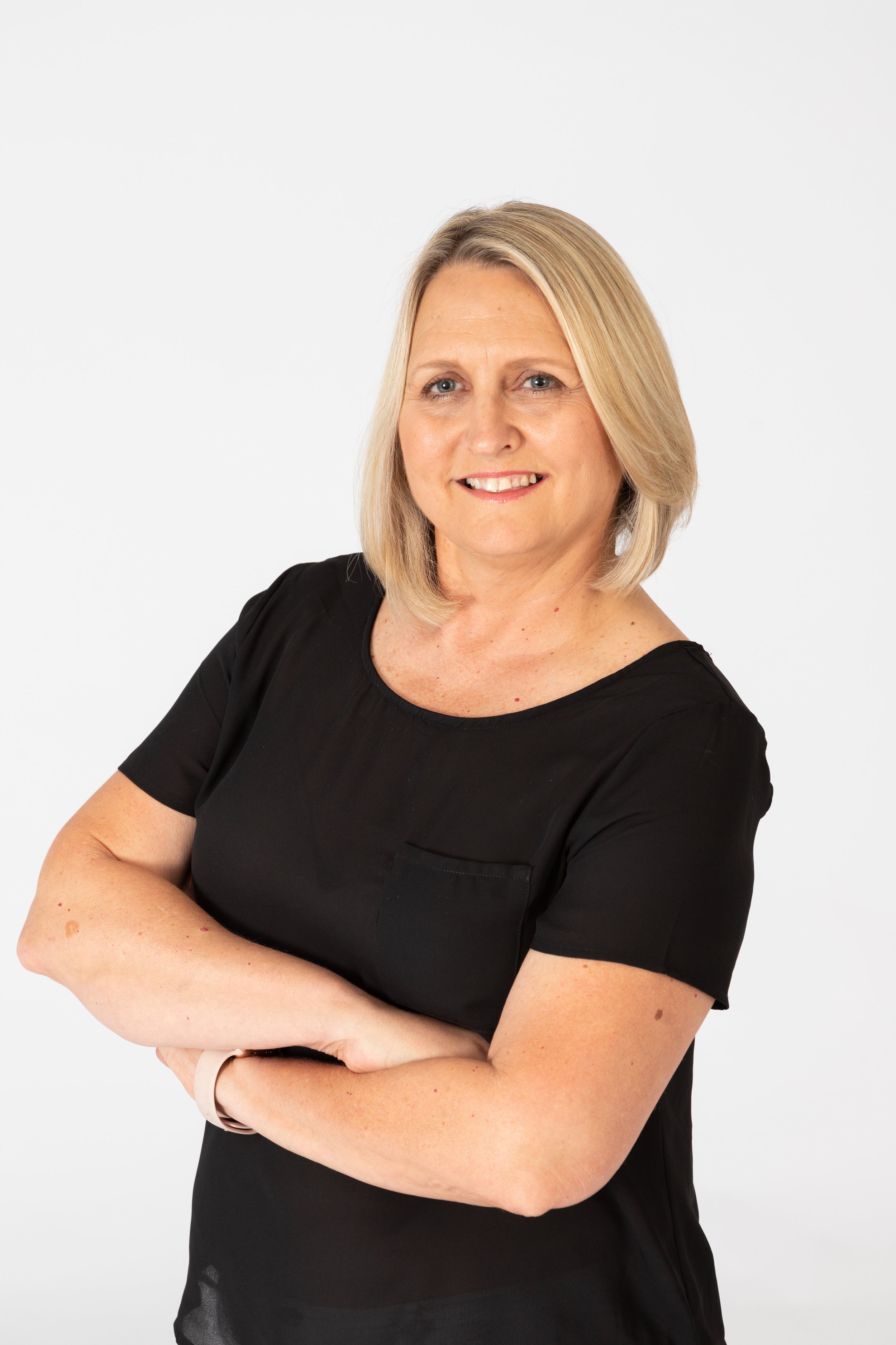 A middle-aged woman with shoulder-length blonde hair, wearing a black shirt, standing with arms crossed against a plain white background.