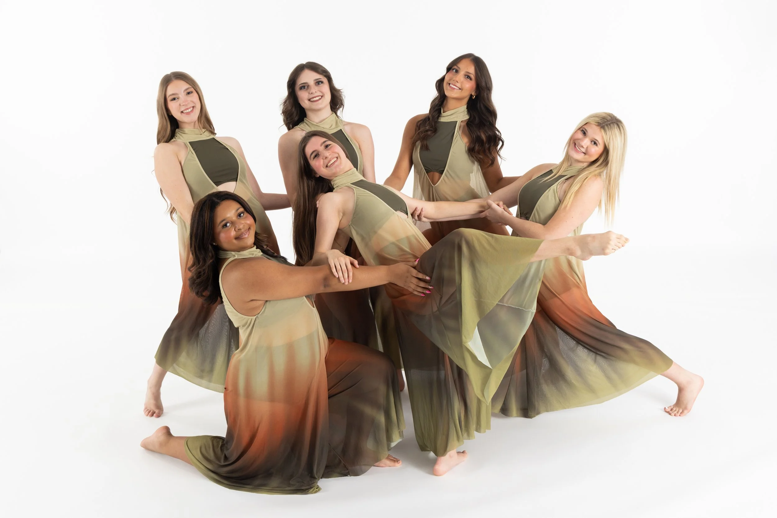 Six women in matching flowy dresses, some kneeling and some standing, holding hands and smiling in a dance pose against a plain white background.