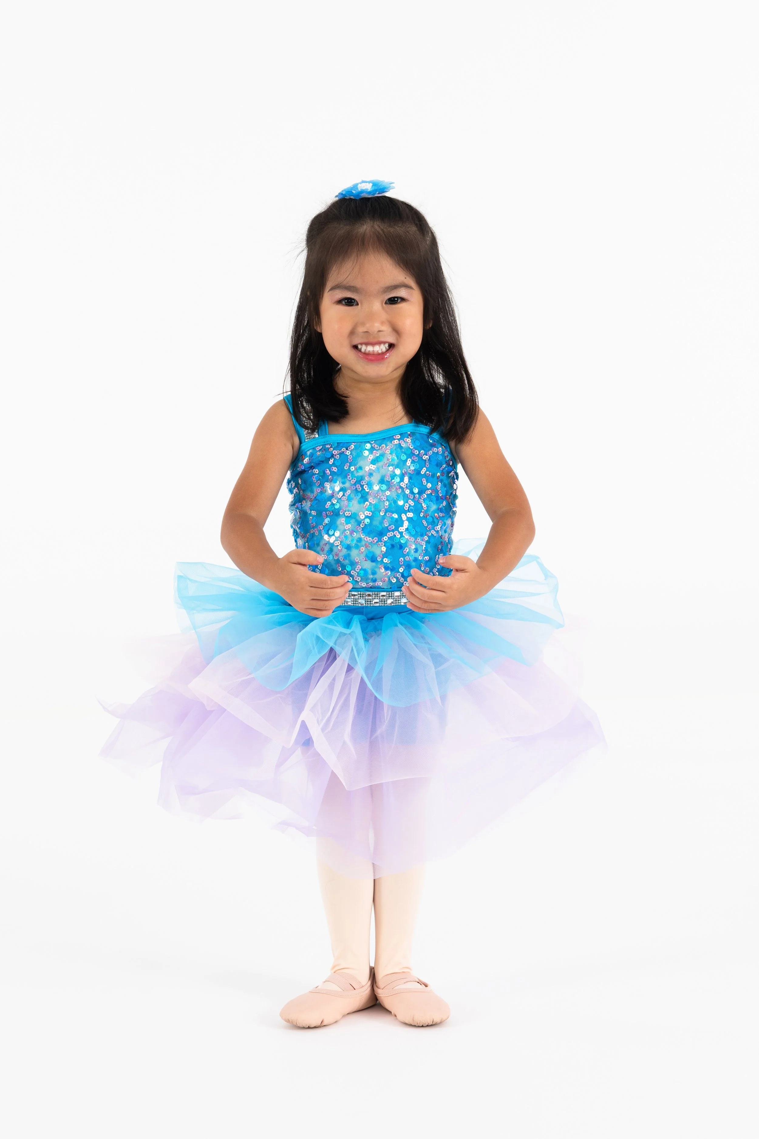 Young girl dressed as a ballerina in a blue, sequined leotard and a pastel-colored tutu, standing against a plain white background.