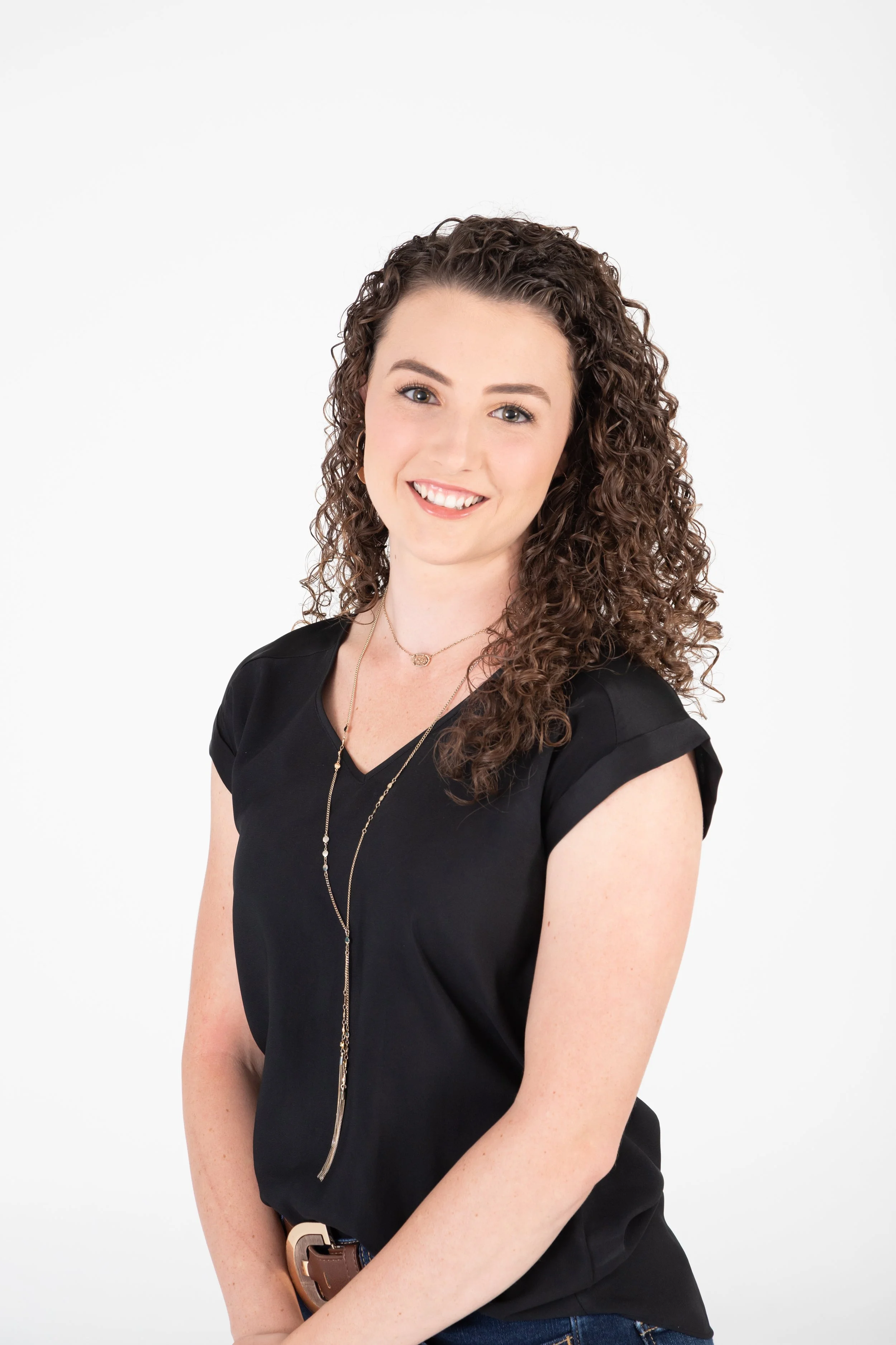 A young woman with curly brown hair smiling at the camera, wearing a black top and layered necklaces, standing against a white background.
