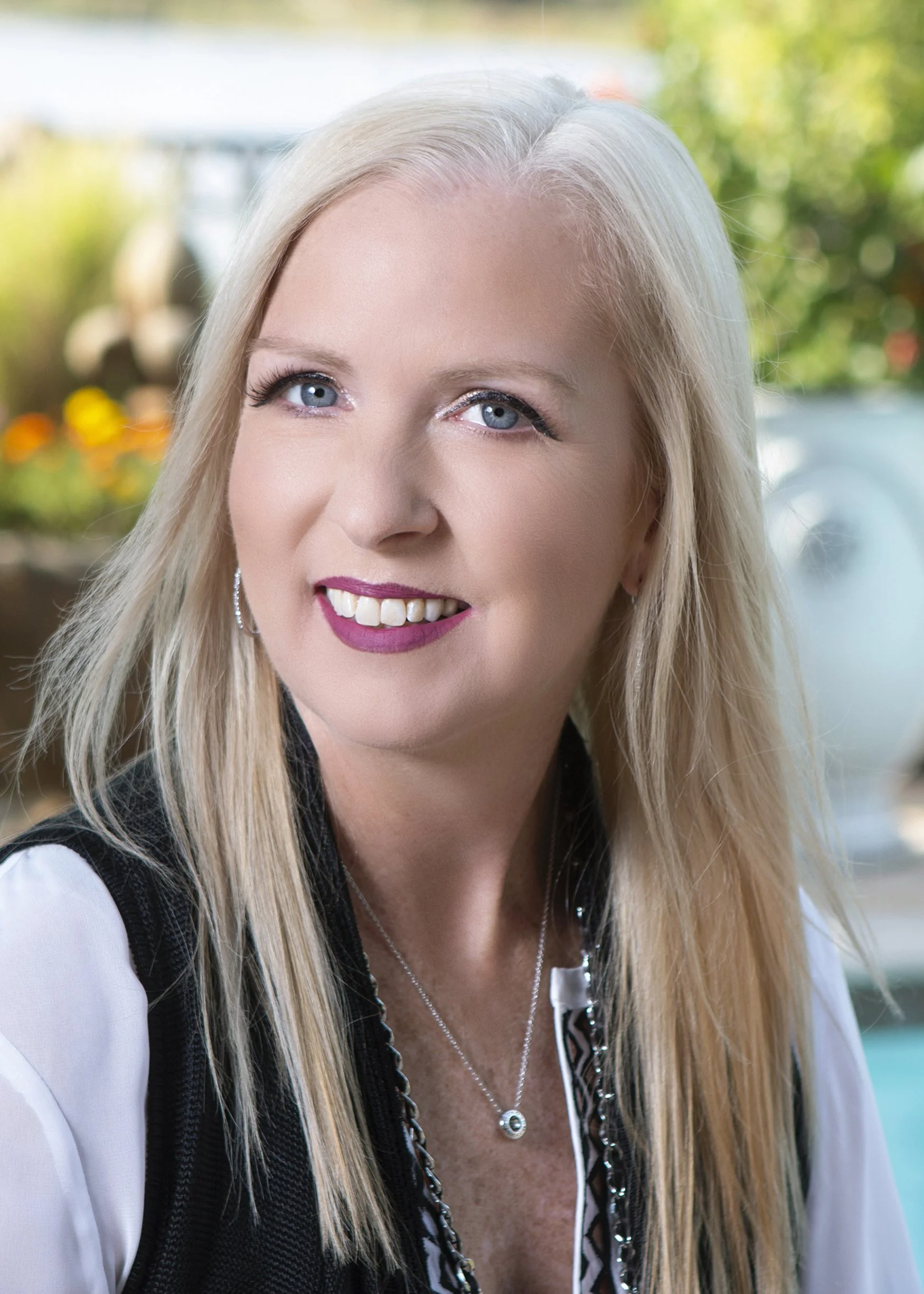 Close-up of a smiling blonde woman with blue eyes, wearing makeup, a silver necklace, earrings, and a black and white shirt with a black vest, outdoors with blurred greenery in the background.