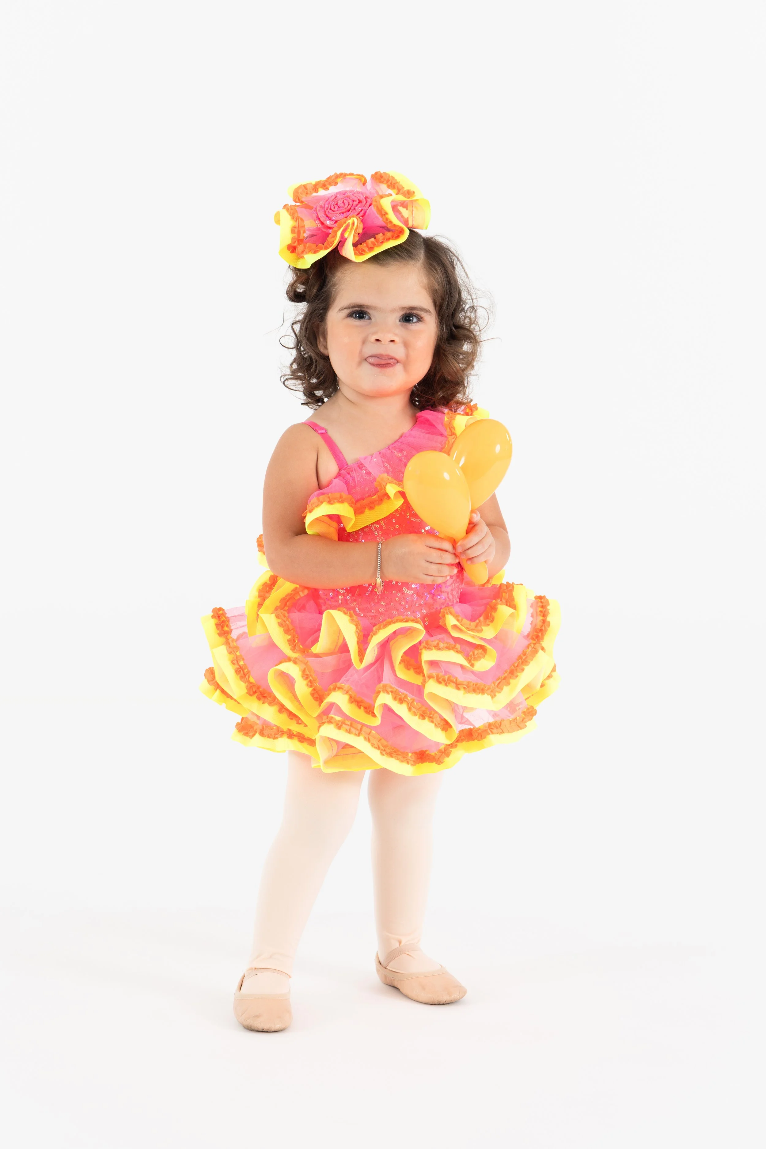 Young girl dressed in bright pink and yellow festive costume holding yellow balloons against a white background.