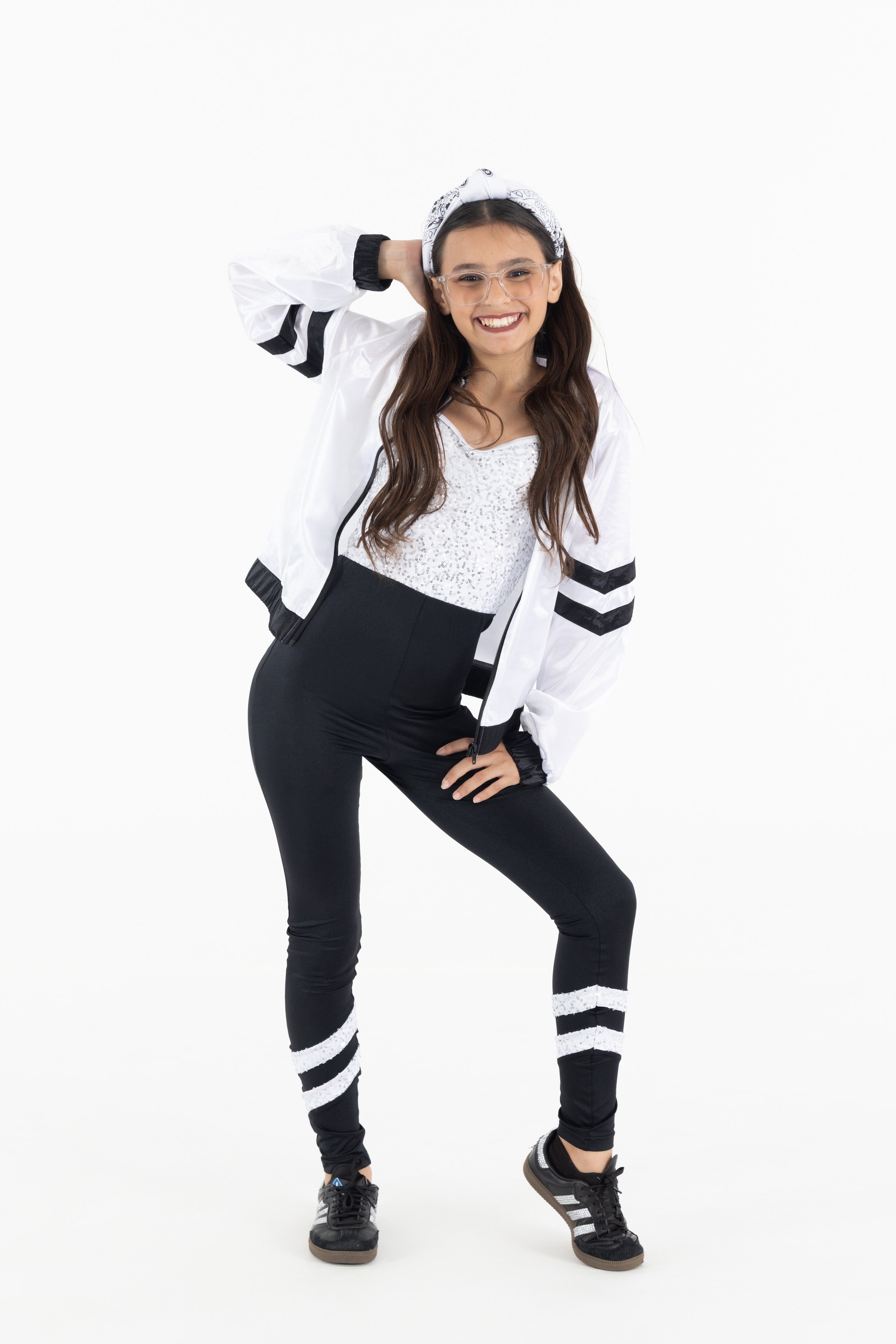 A young girl with long brown hair, wearing clear glasses, a white bandana, a shiny white jacket with black stripes on the sleeves, a white sequin top, black leggings with white stripes on the calves, and black sneakers, poses with one hand on her hip and the other behind her head against a plain white background.