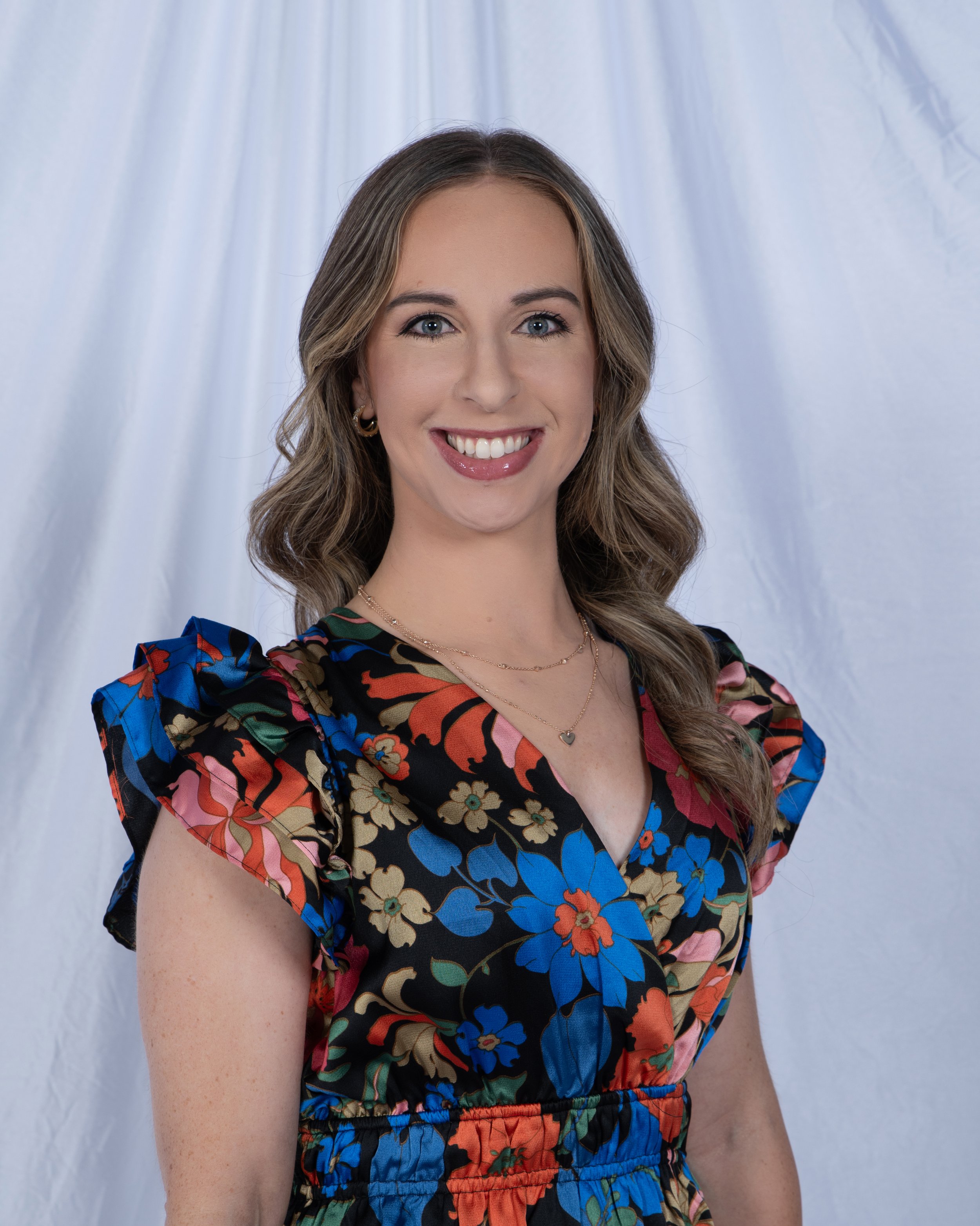 A woman with light brown hair smiling, wearing a colorful floral dress with short sleeves, and a gold necklace, standing against a white fabric backdrop.