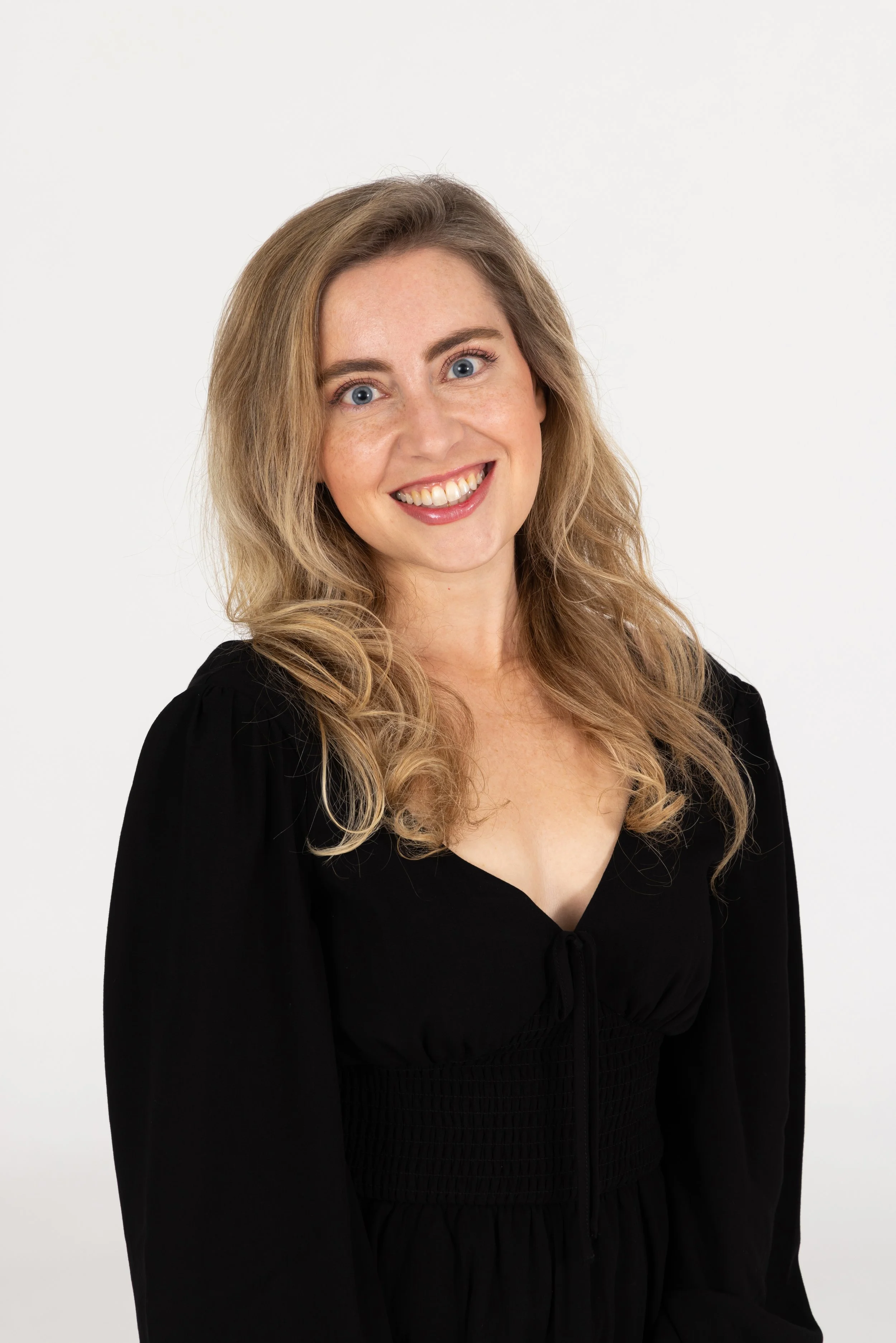 Portrait of a young woman with wavy blonde hair, blue eyes, smiling, wearing a black top, against a plain white background.
