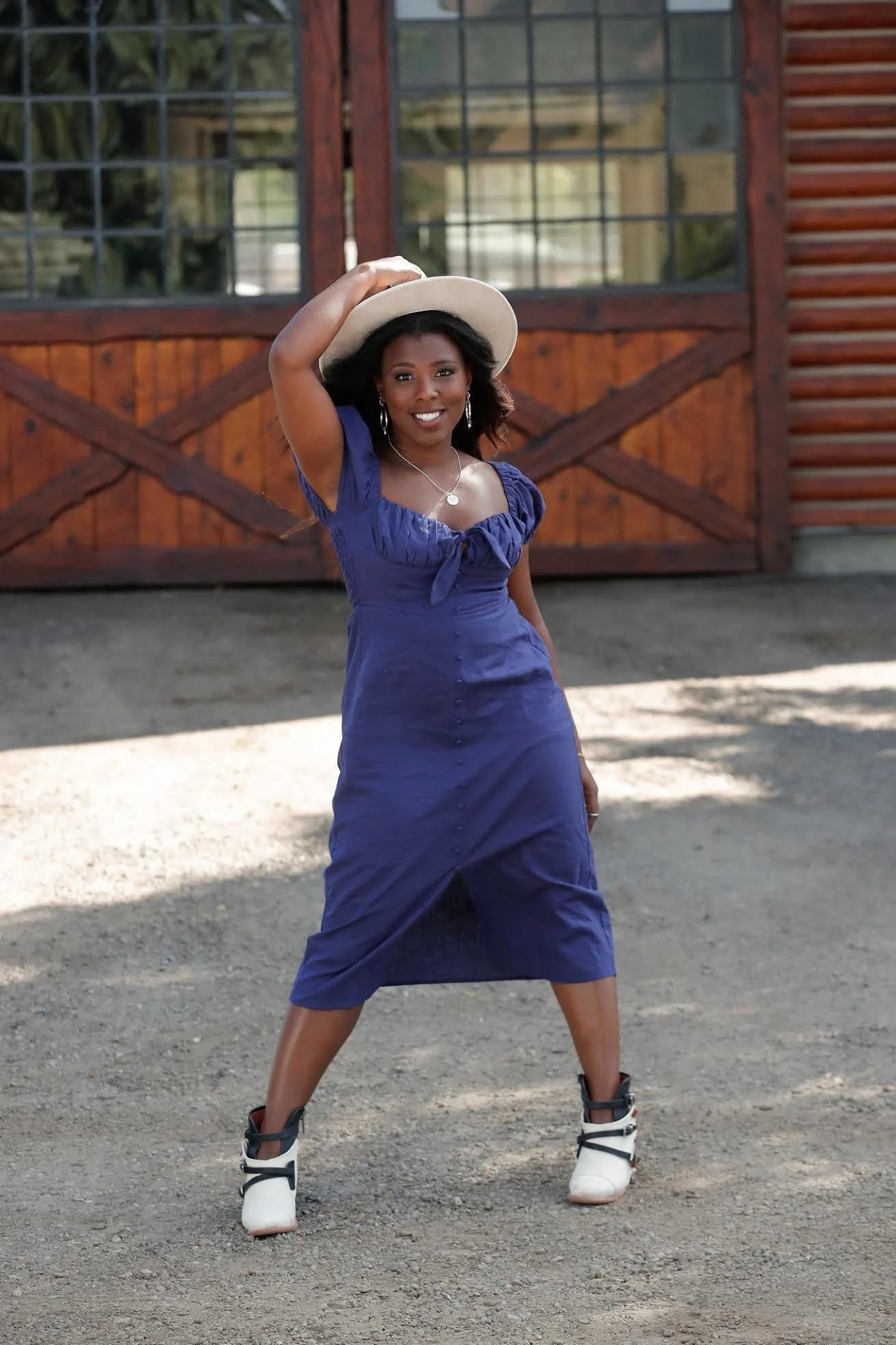 A woman in a blue dress and white boots standing in front of a wooden barn, smiling, with her hand on her hat.