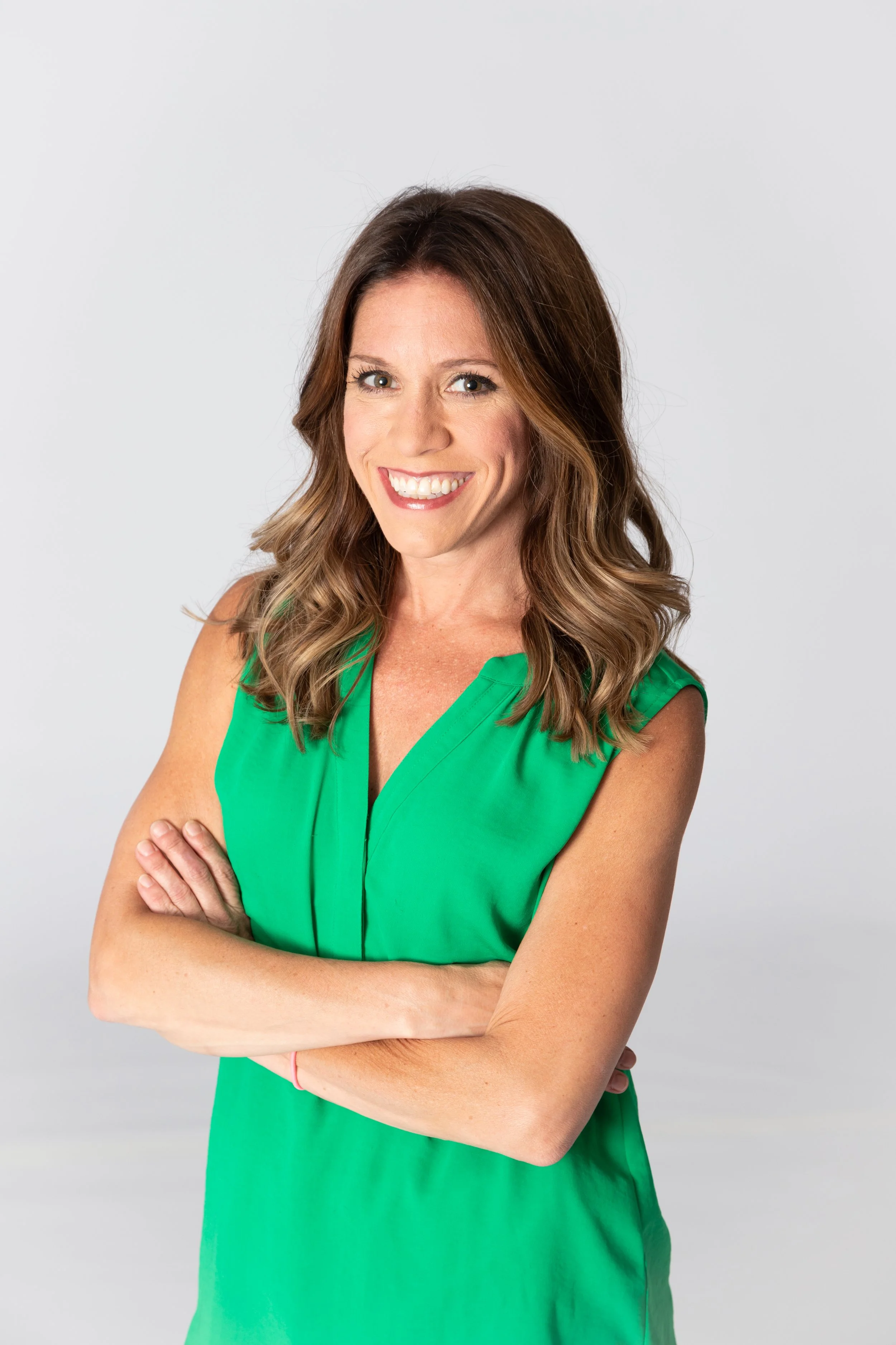 A smiling woman with long, wavy brown hair, wearing a sleeveless green dress, standing with arms crossed against a plain light gray background.