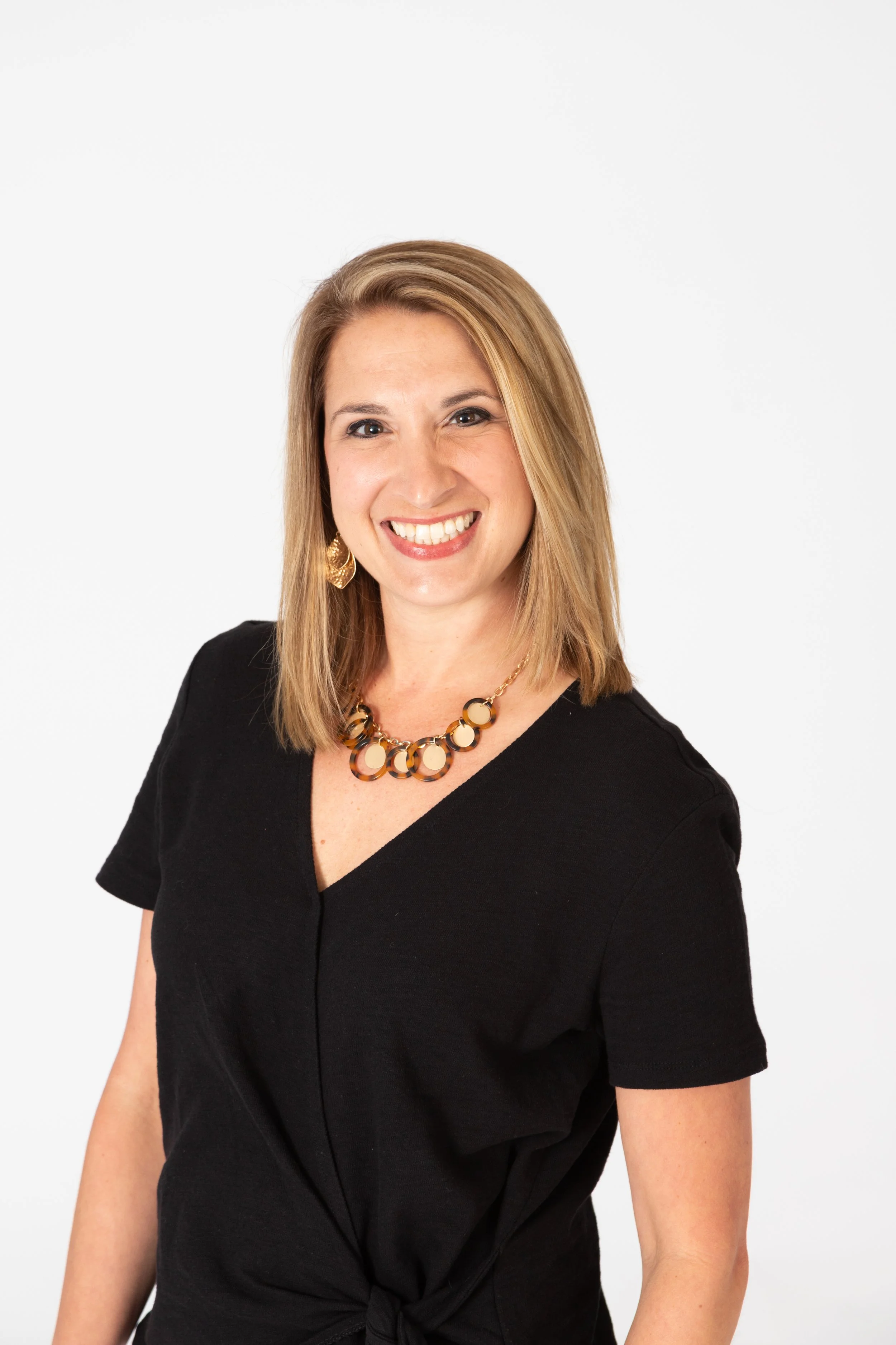 A woman with shoulder-length blonde hair smiling, wearing gold earrings and a black dress with a tie knot at the waist, against a white background.