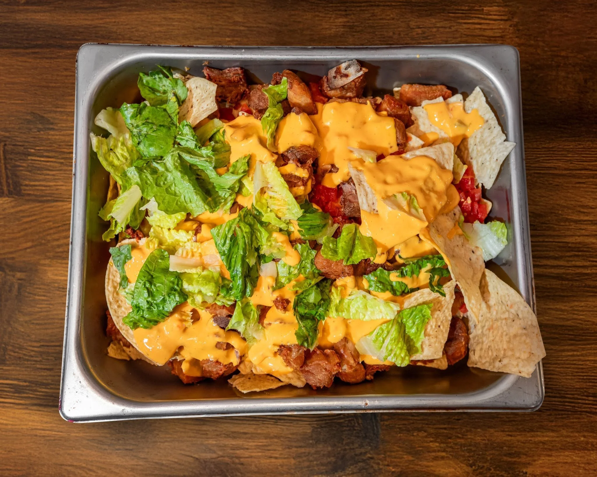 A metal tray filled with nachos, topped with shredded lettuce, melted cheese, ground beef, and chopped tomatoes, sitting on a wooden table.