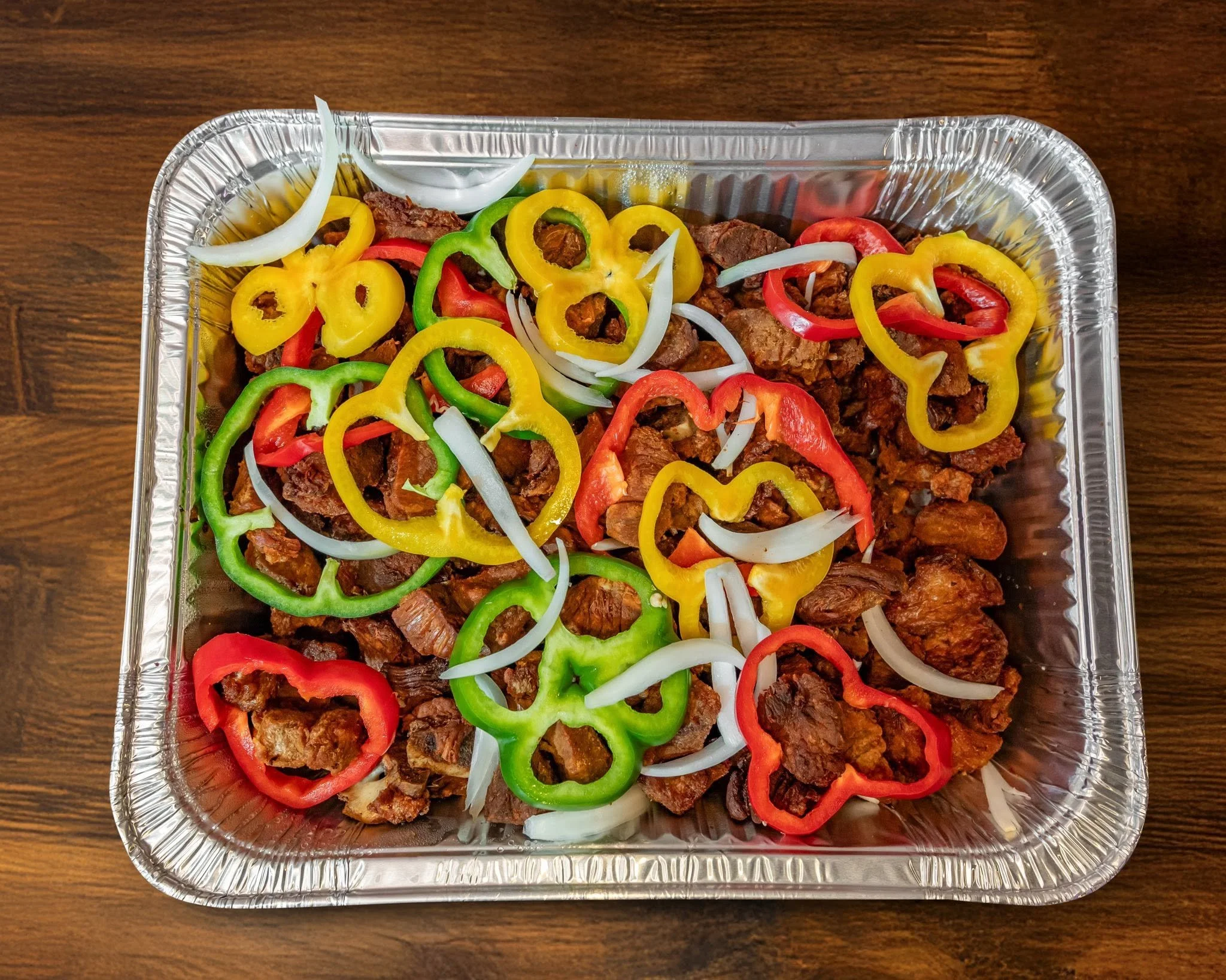 Aluminum takeout container filled with cooked beef chunks topped with sliced red, yellow, green, and white bell peppers and onions, placed on a wooden surface.