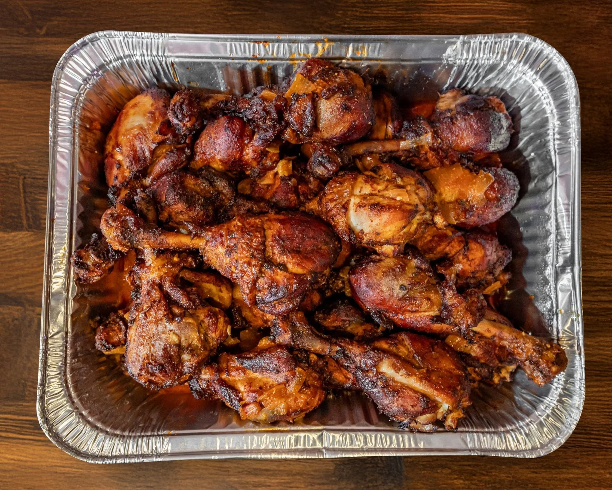Aluminum tray filled with cooked chicken legs and pieces in a dark sauce on a wooden surface.