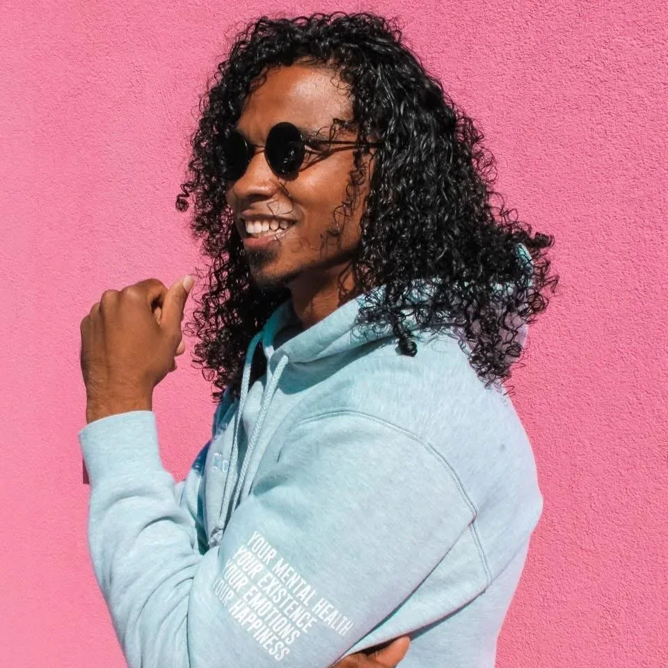 A man with curly black hair wearing sunglasses and a light blue hoodie with white text, smiling against a pink wall.