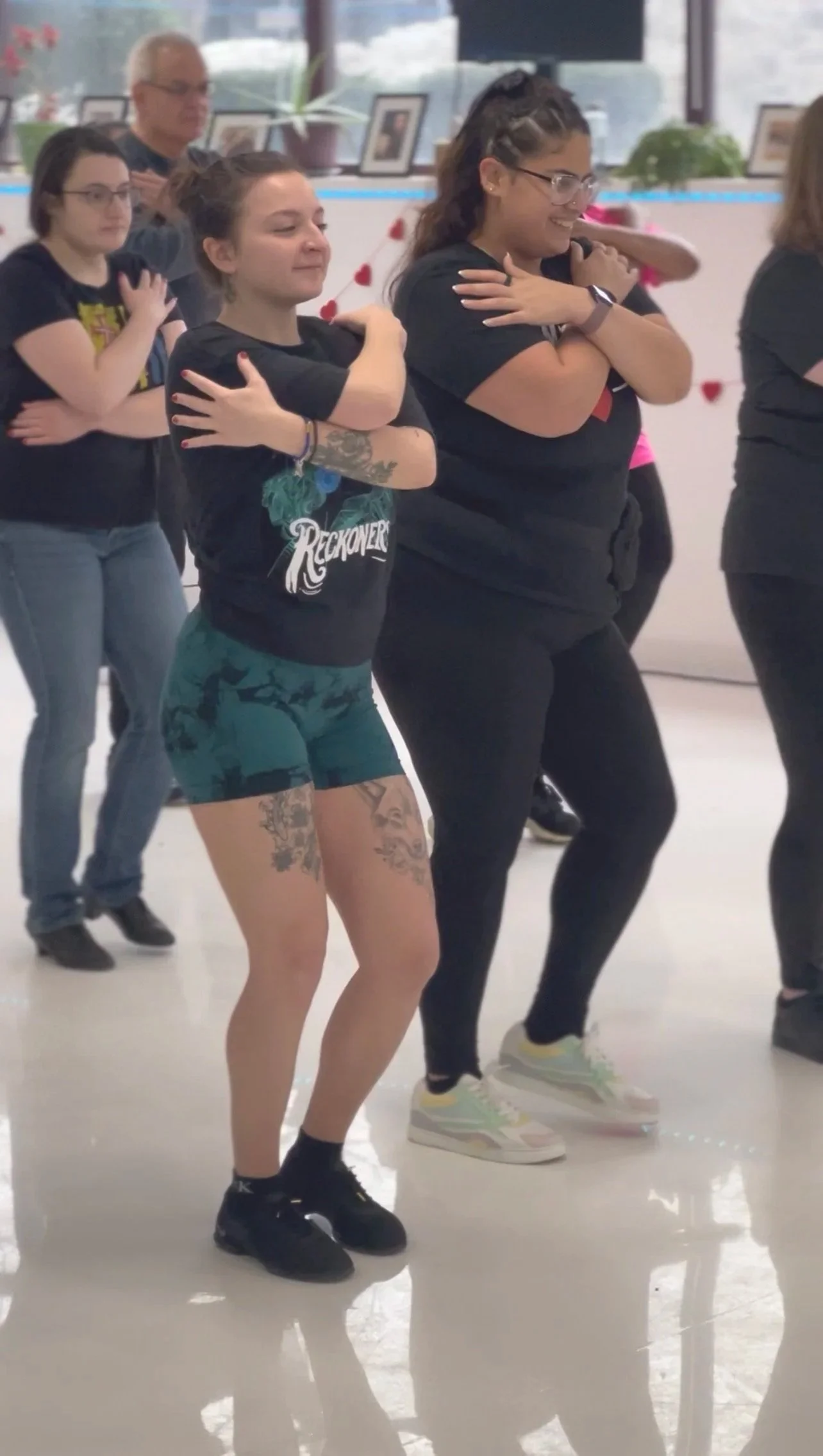 Group of people practicing a choreography or dance in a bright indoor studio, with some crossing their arms over their chests and smiling.