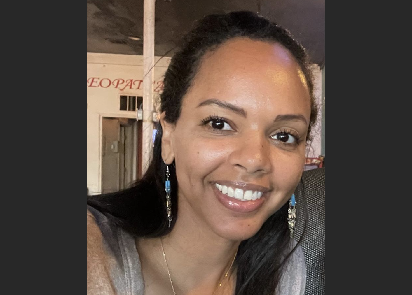 A smiling woman with dark hair, earrings, and a necklace, sitting indoors in a casual setting with a background that includes a partially visible sign and a doorway.