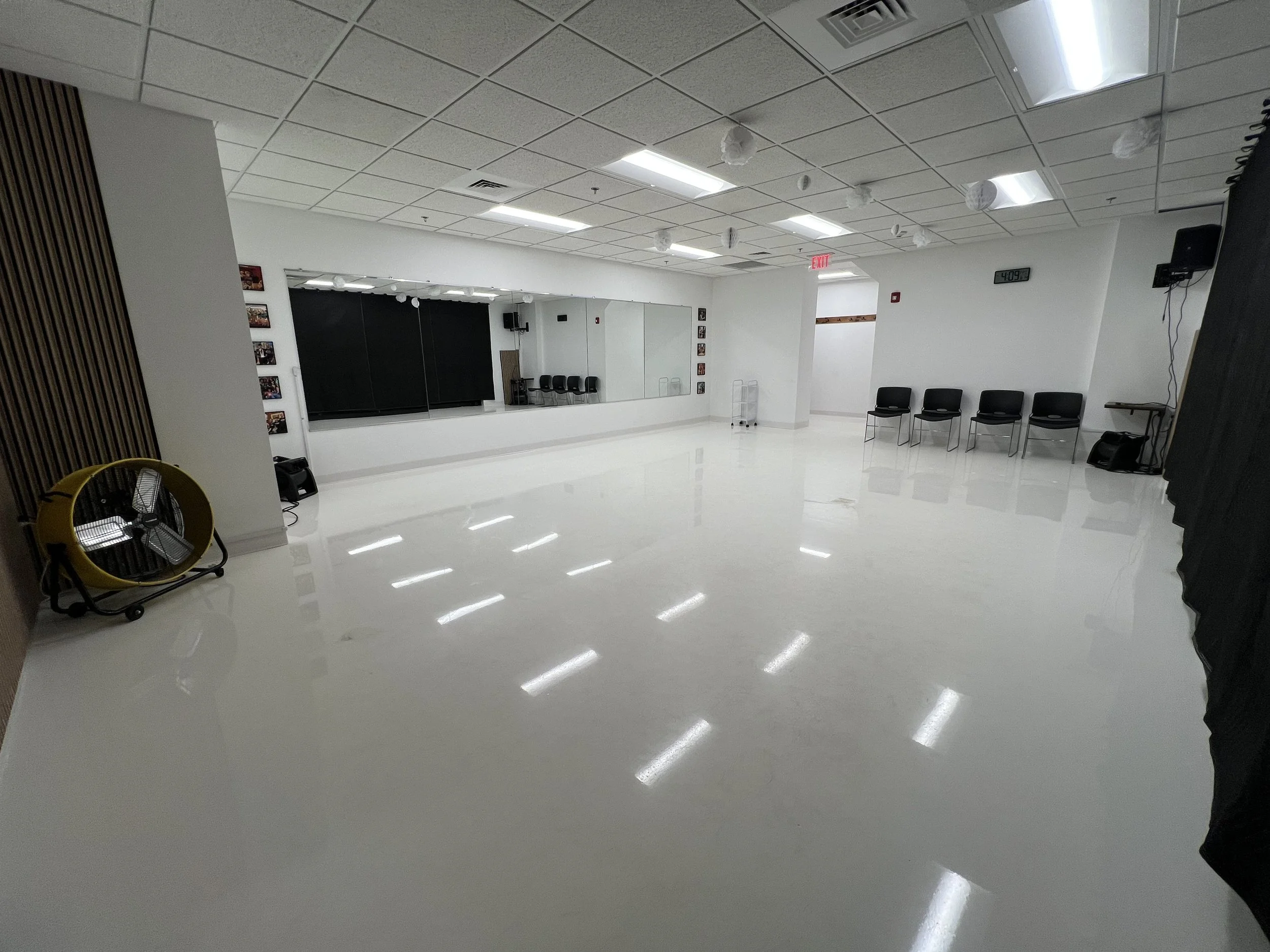 An empty room with white walls, a white tiled ceiling, and a shiny white floor. There are several chairs along the walls, a large mirror, and black curtains. The room has fluorescent lighting and a digital clock on the wall showing 4:09.