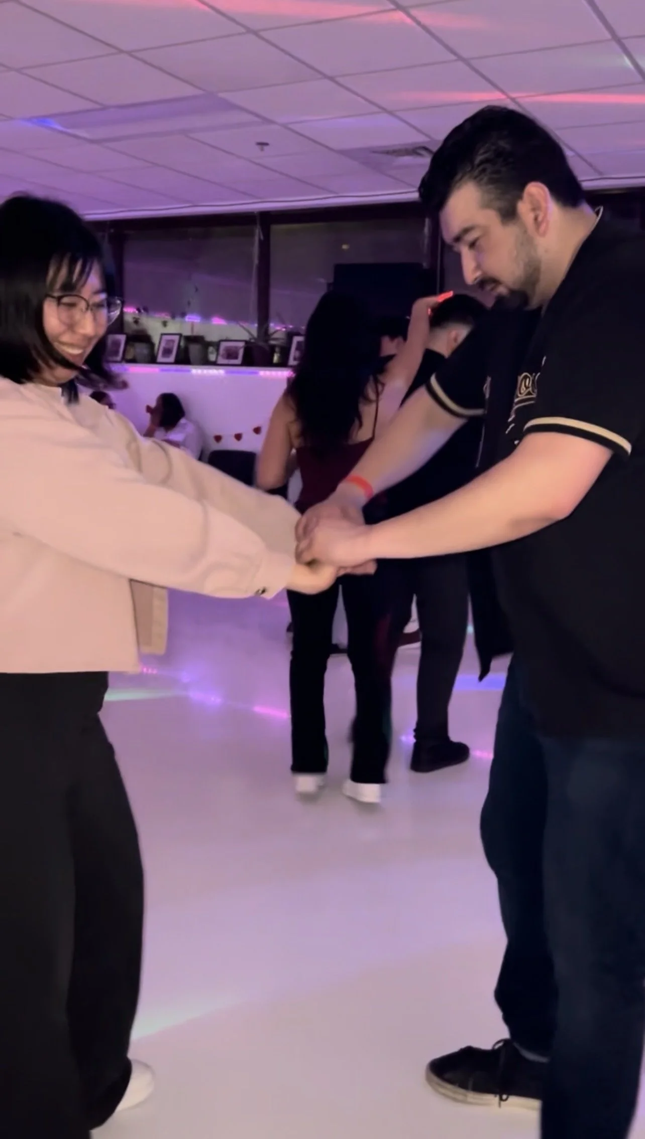 Two people holding hands and dancing together in a party setting with purple and pink lighting, with other people in the background.