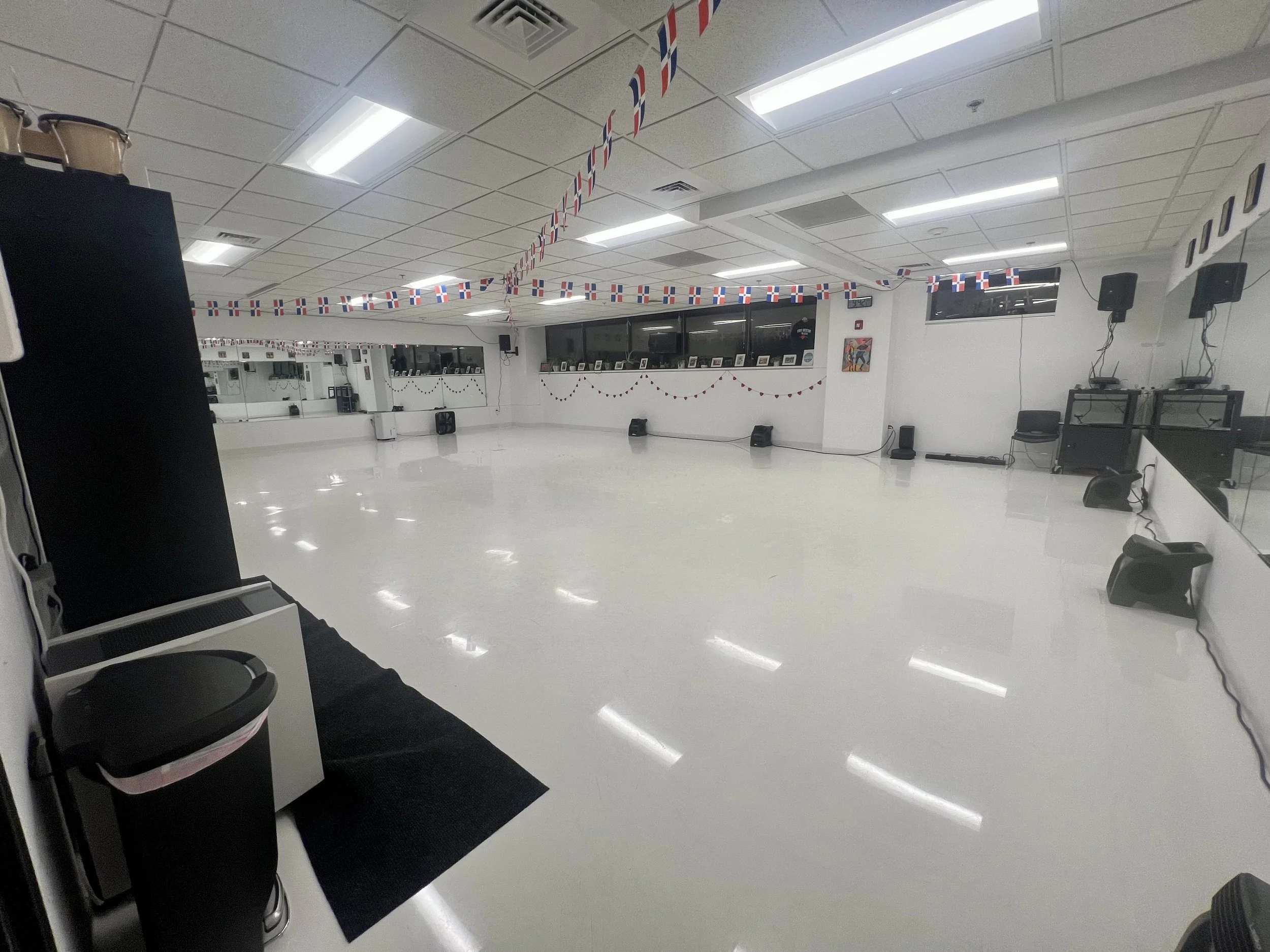 Dancer or instructor in a dance studio with American flag decorations hanging from the ceiling. The room has a mirror wall, fluorescent lighting, and audio speakers.