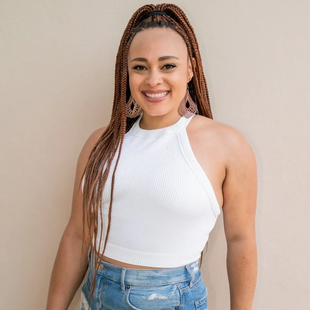 A woman with long braided hair smiling, wearing a white sleeveless top and denim shorts, standing against a beige wall.
