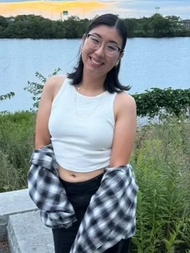 A young woman with dark hair, glasses, and a white sleeveless top smiling outdoors near a body of water with greenery in the background.