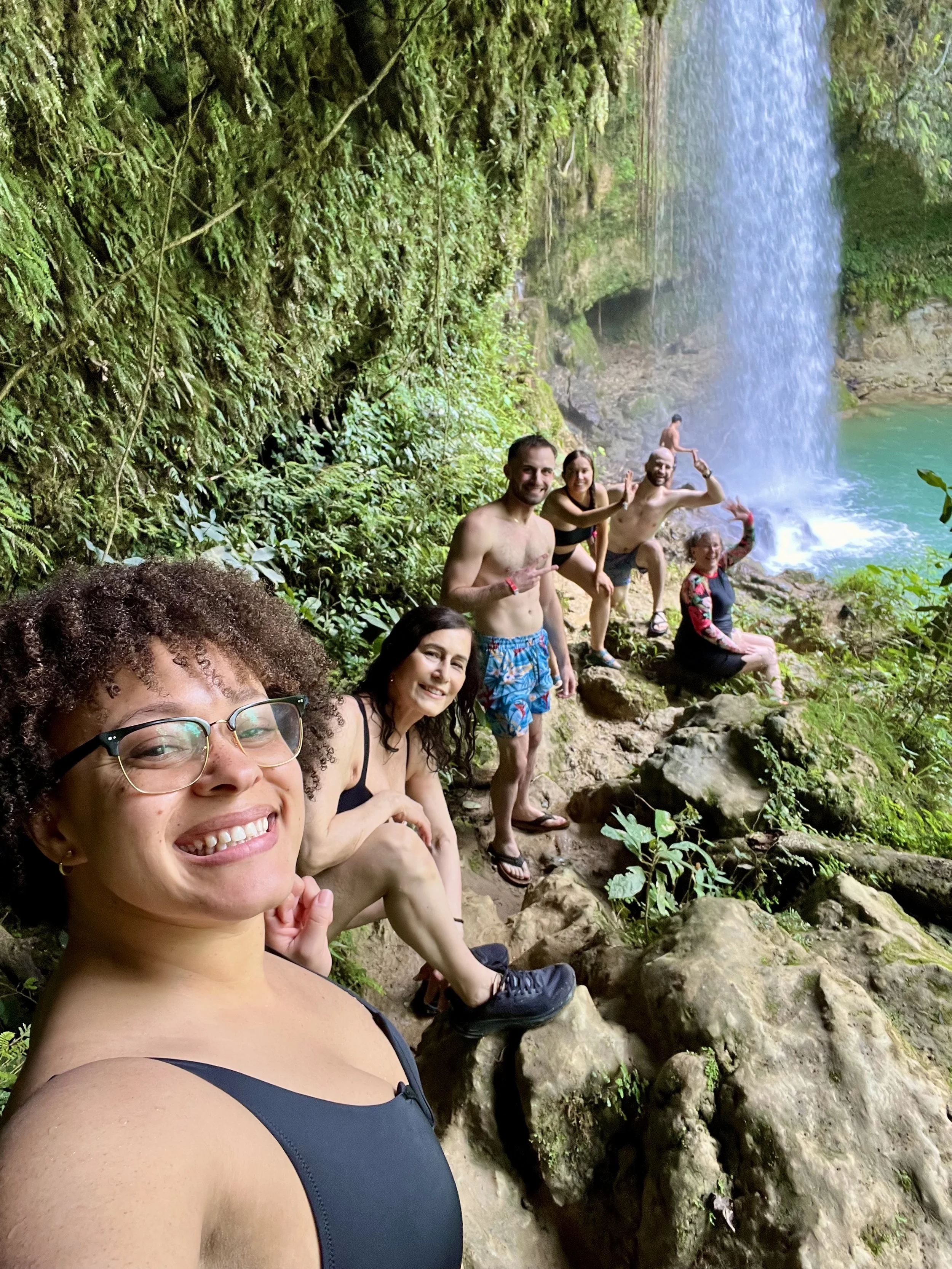 A group of smiling people sitting and standing along a rocky path near a waterfall in a lush green forest.