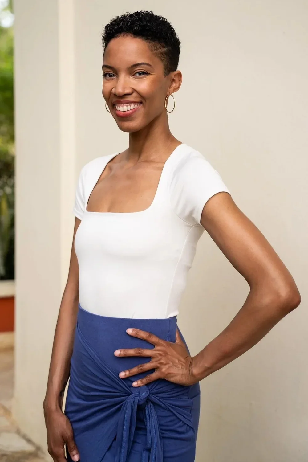 Portrait of a smiling woman with short curly hair, wearing hoop earrings, a white top, and a blue wrap skirt tied at her waist, standing indoors.