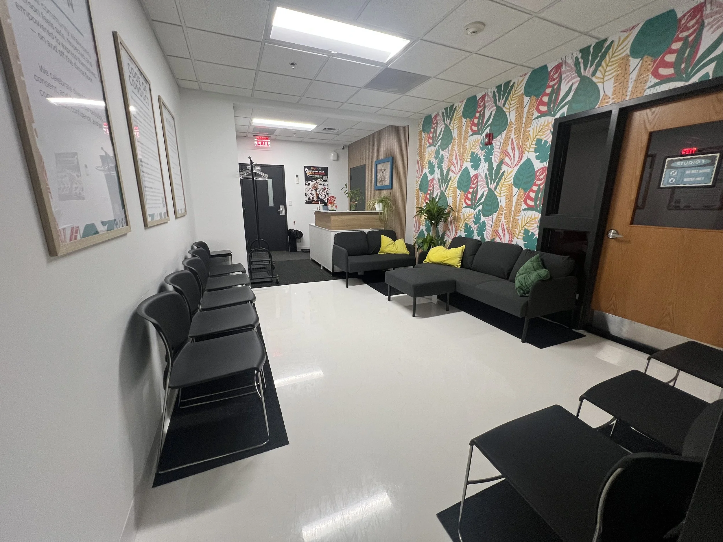 A waiting room with black chairs lined up against the left wall, a sofa with yellow and green pillows on the right, a small table with potted plants, and a vibrant tropical leaf wallpaper on one wall. The ceiling has fluorescent lighting and an emergency exit sign.