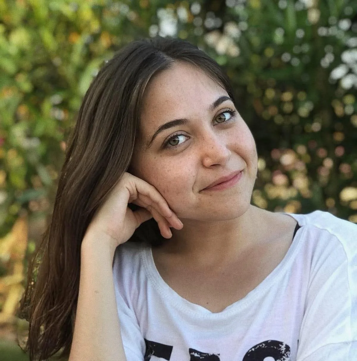 A young woman with long brown hair, light skin, and gray eyes, smiling softly and resting her chin on her hand, outdoors with a blurred background of greenery and flowers.